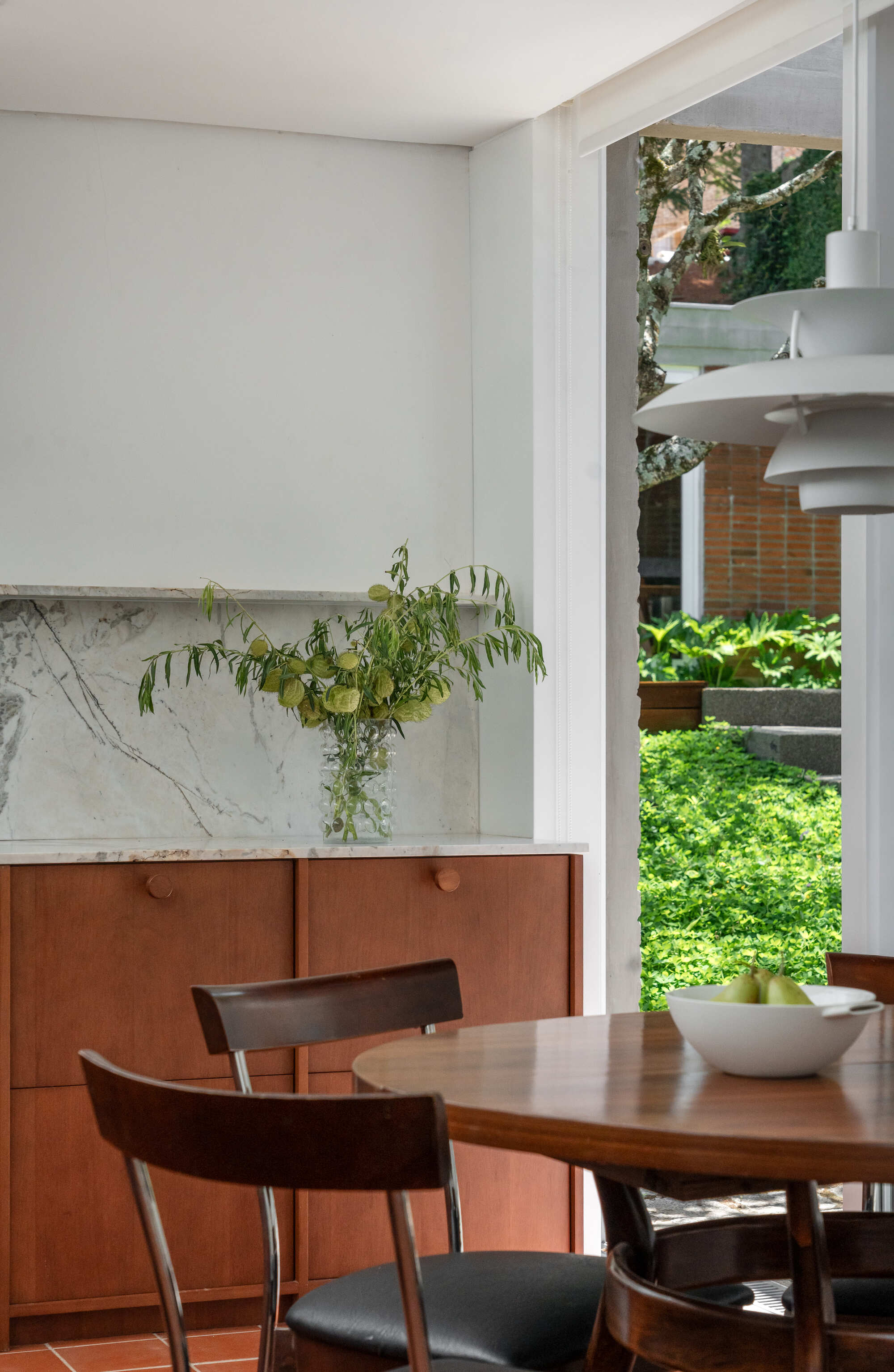 Timber cabinet with marble backsplash and a vase of green stems beside a round table, opening to a sunlit garden