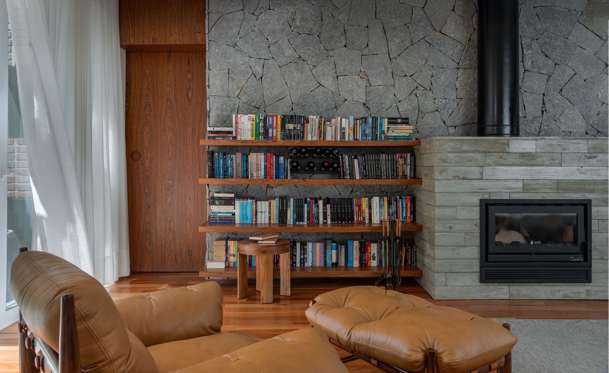 Built-in bookshelves against a dry-laid grey stone wall beside a concrete-clad fireplace, with leather armchair and ottoman in the foreground