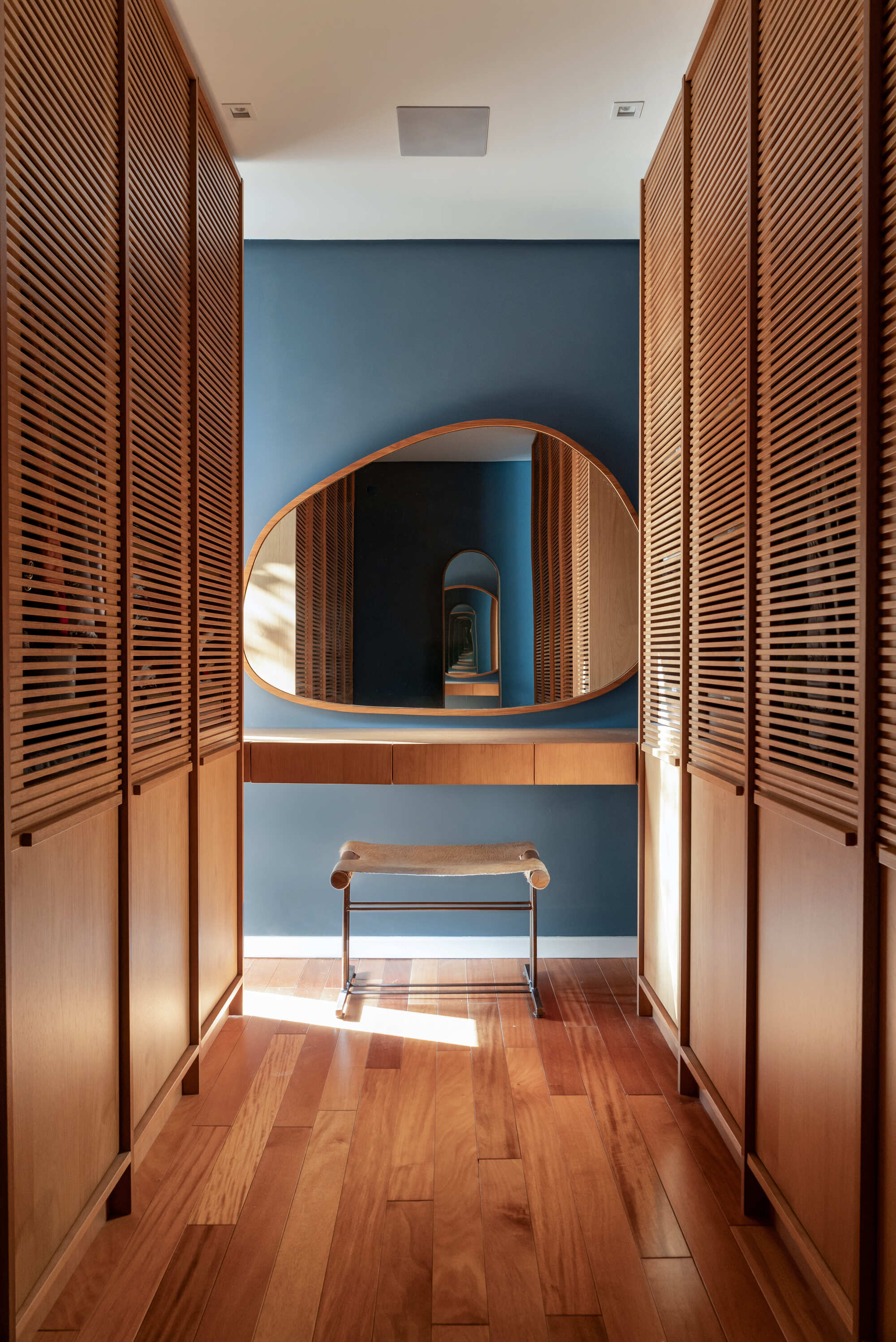 Narrow dressing corridor lined with slatted timber wardrobe doors, ending in a pebble-shaped mirror over a blue wall, with a sunbeam on the floor