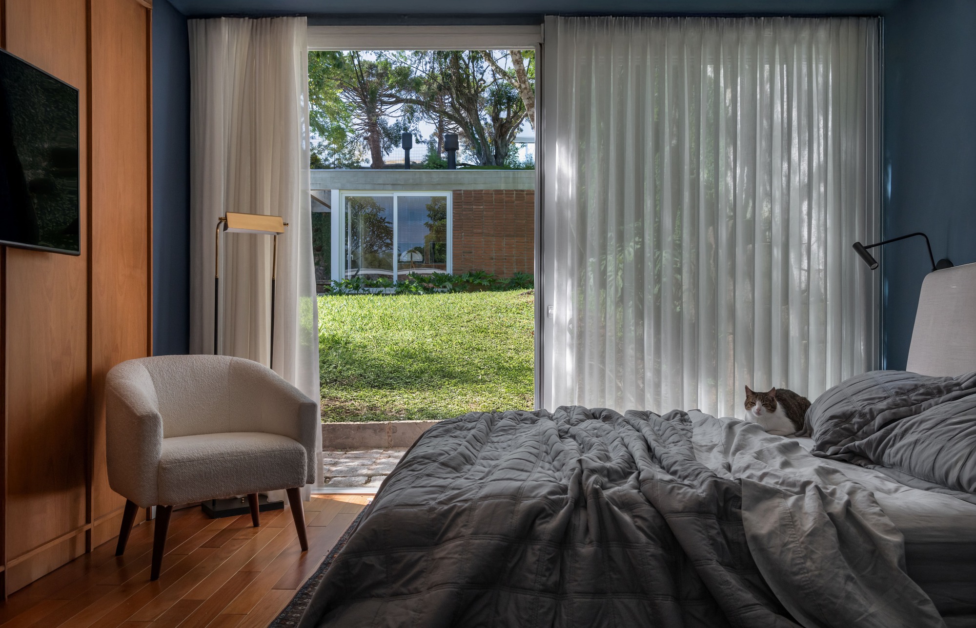Bedroom with sheer curtains framing a sliding glass door onto a lawn, a cat resting on the grey bedding