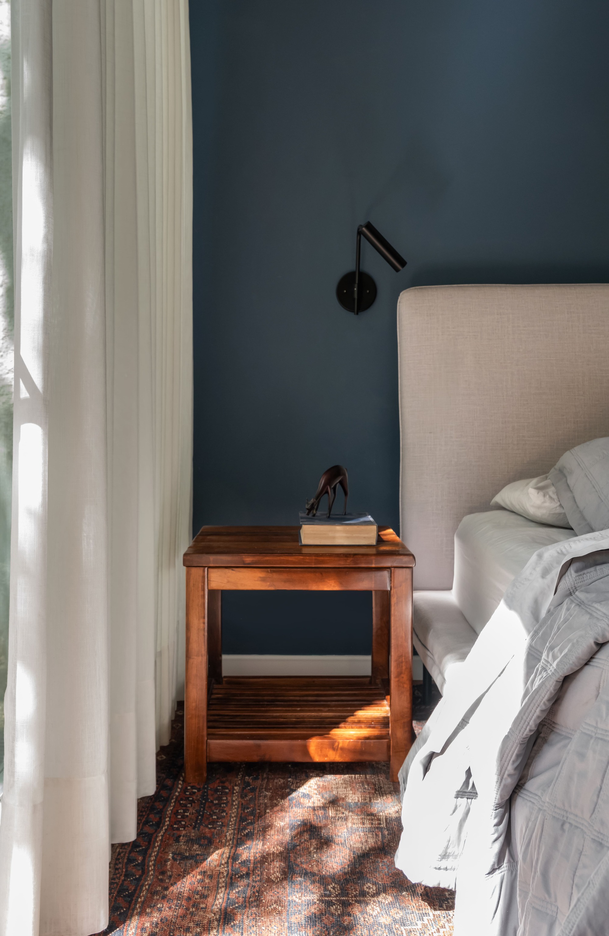 Wooden bedside table with a book and small figurine under a black wall lamp, dappled sunlight falling across a patterned rug