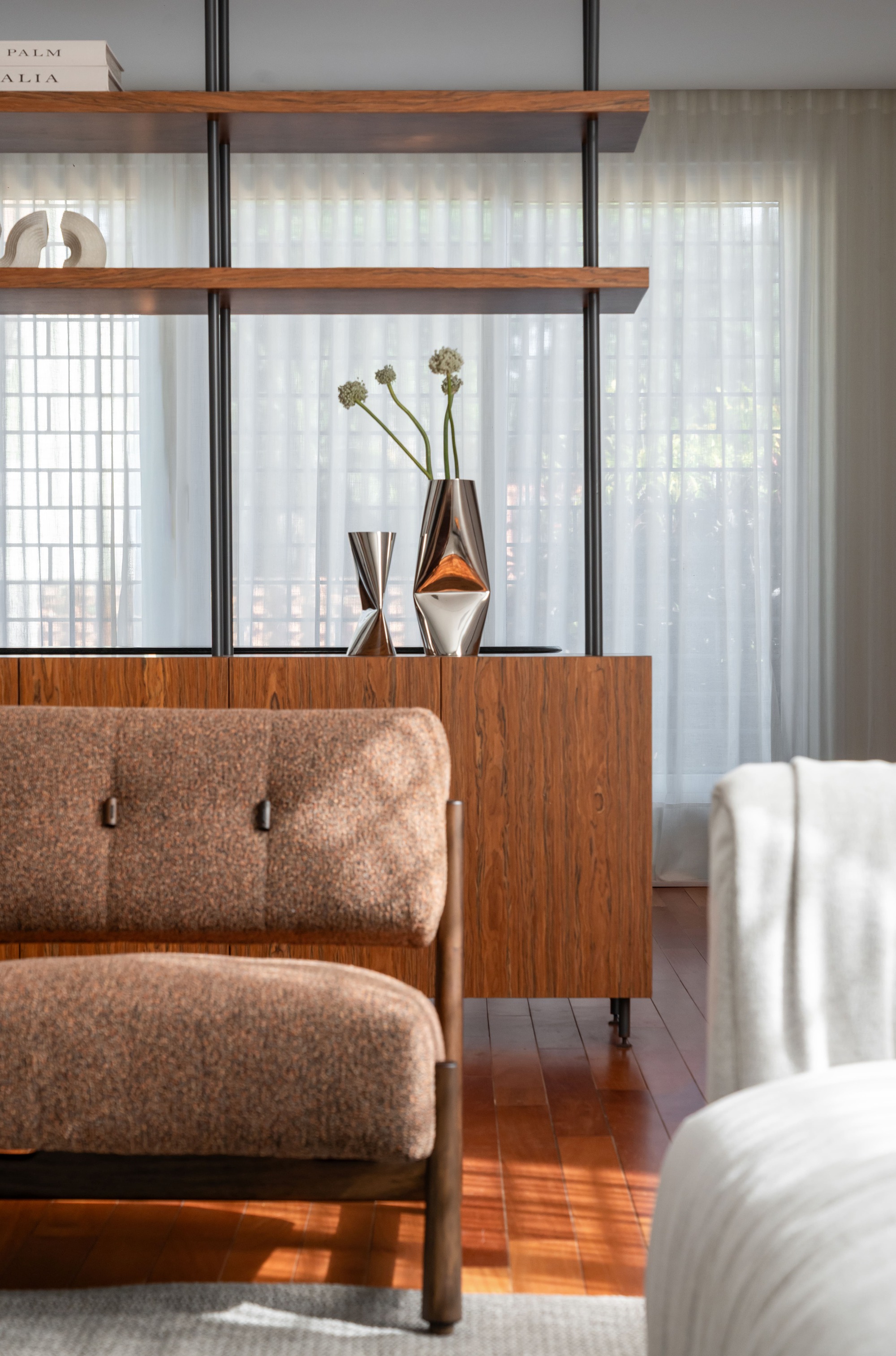 Walnut sideboard with metallic vases and open shelving in front of sheer curtains, a bouclé armchair in the foreground catching afternoon light