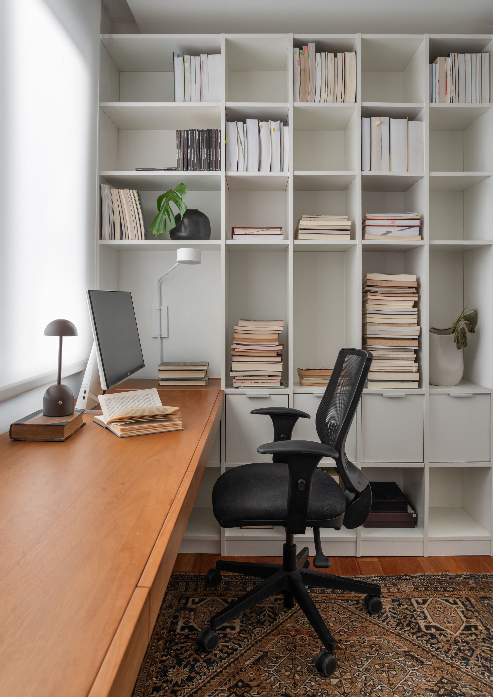 Home office with a long timber desk, white cubby shelving full of books and a black mesh chair on a patterned rug, lit by a frosted window