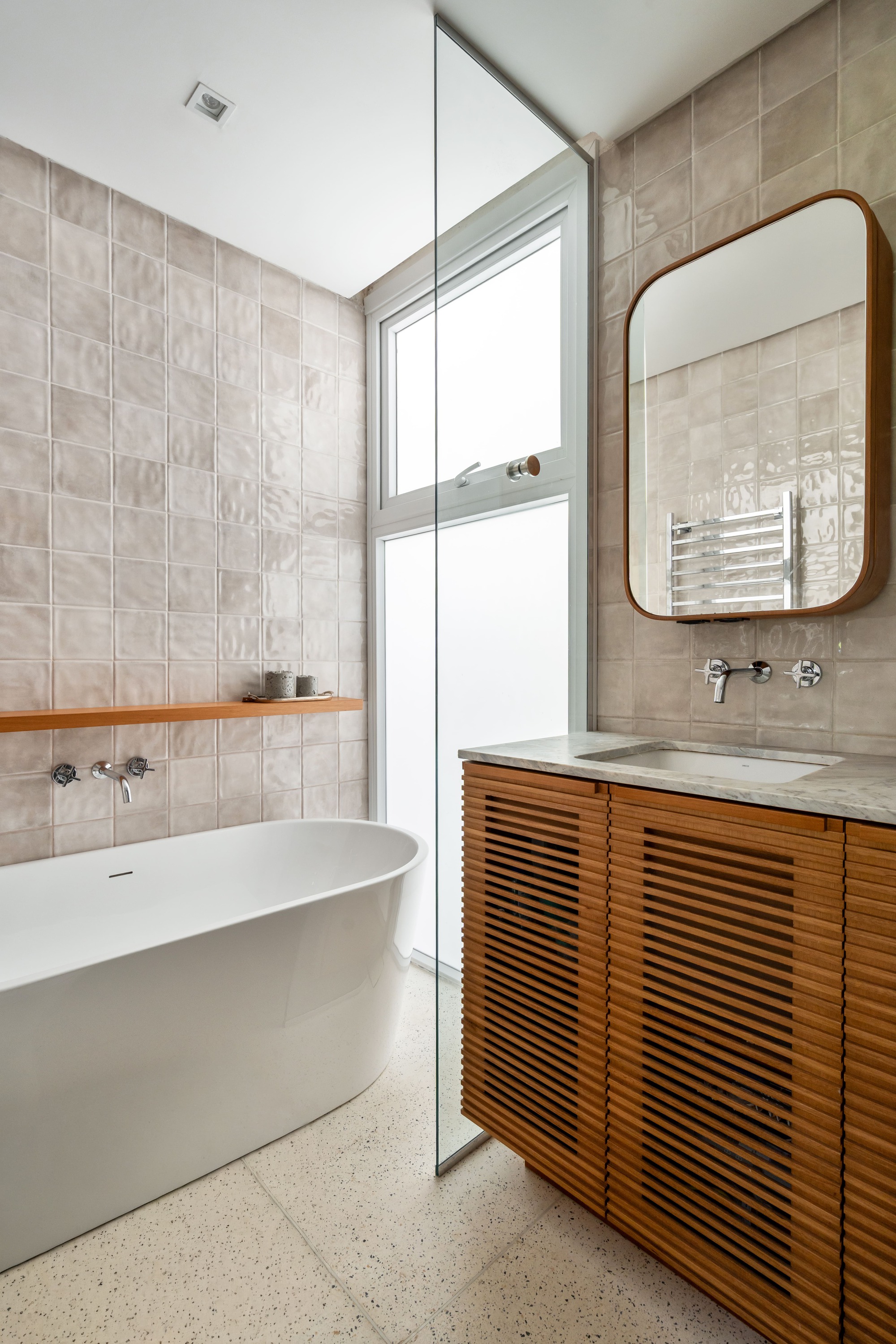 Bathroom with a freestanding white tub, glazed beige tile walls and a slatted timber vanity beside a frosted window in soft daylight