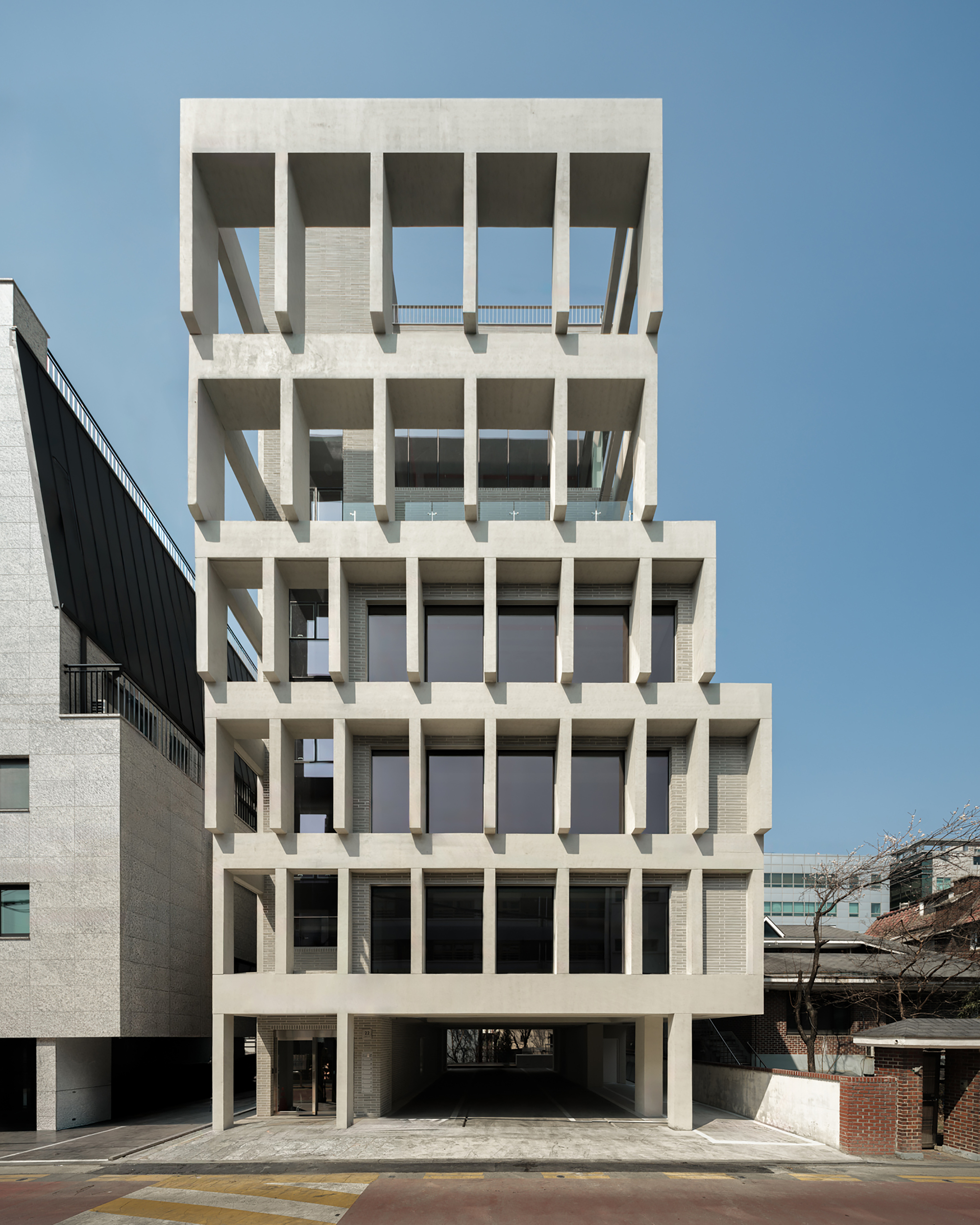 Frontal view of the stepped concrete grid facade with deep fins and a open ground-floor passage under a clear blue sky