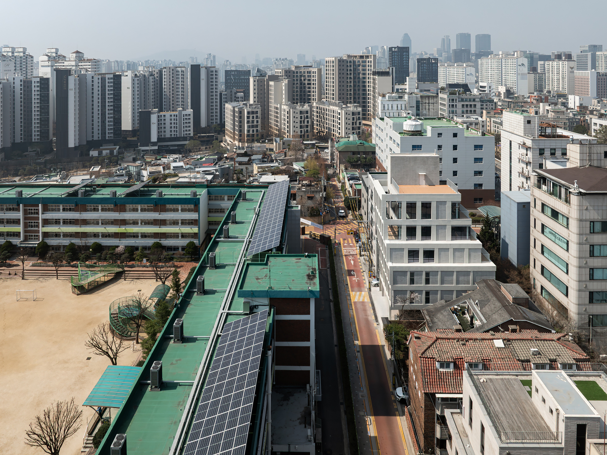 Elevated view over low rooftops with solar panels toward the pale tiered building beside a narrow street in hazy light