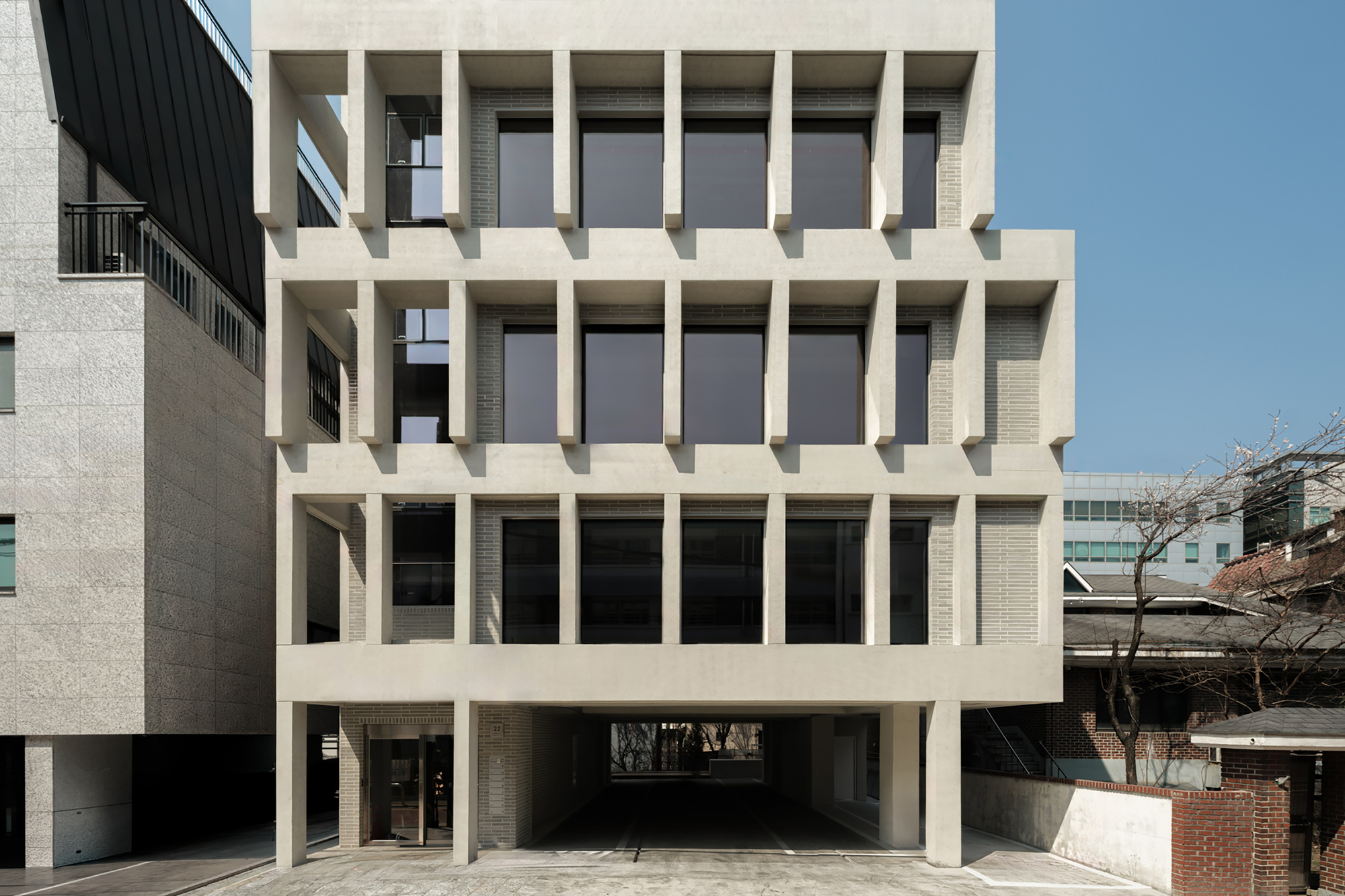 Facade elevation showing vertical concrete fins over dark windows and brick panels, with a bare tree at right