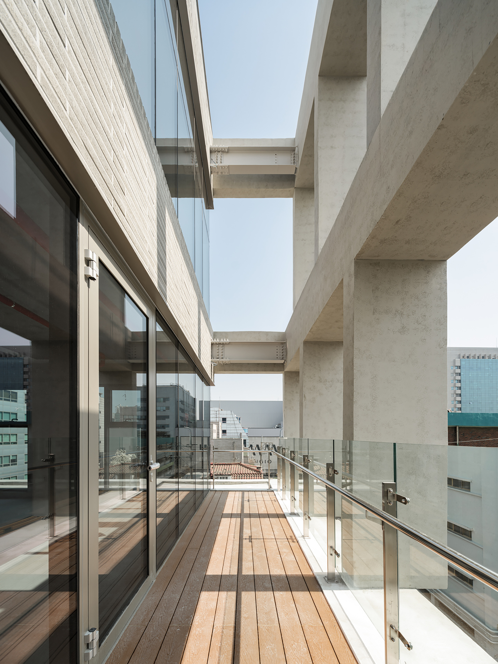 Timber-decked terrace between a glazed brick wall and concrete frame, with a glass balustrade in bright sunlight