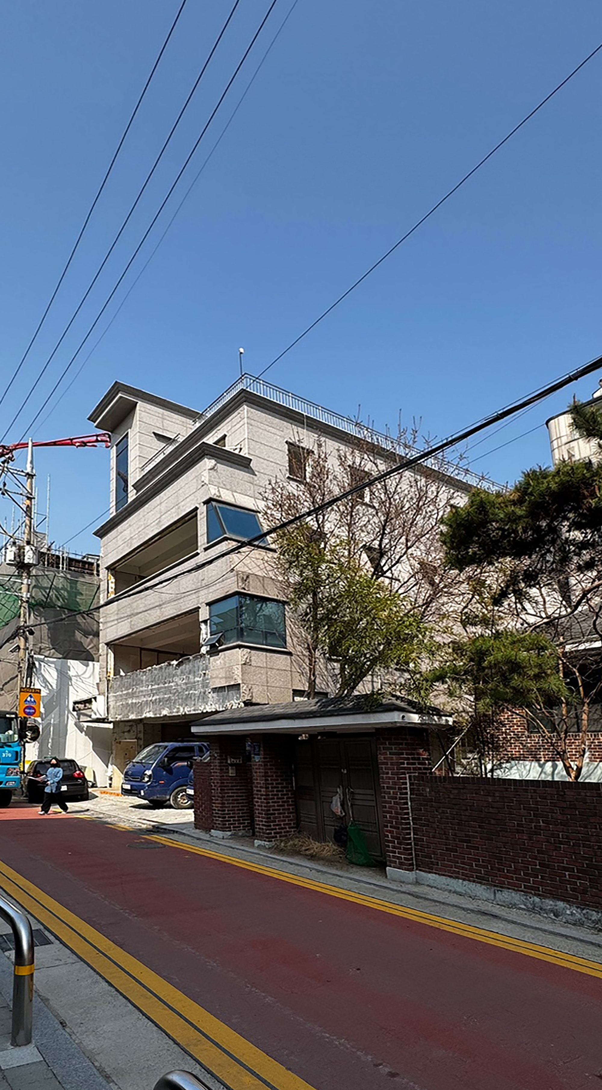 Street view of the stone-clad building with recessed terraces behind a brick wall and gate, under power lines and clear blue sky