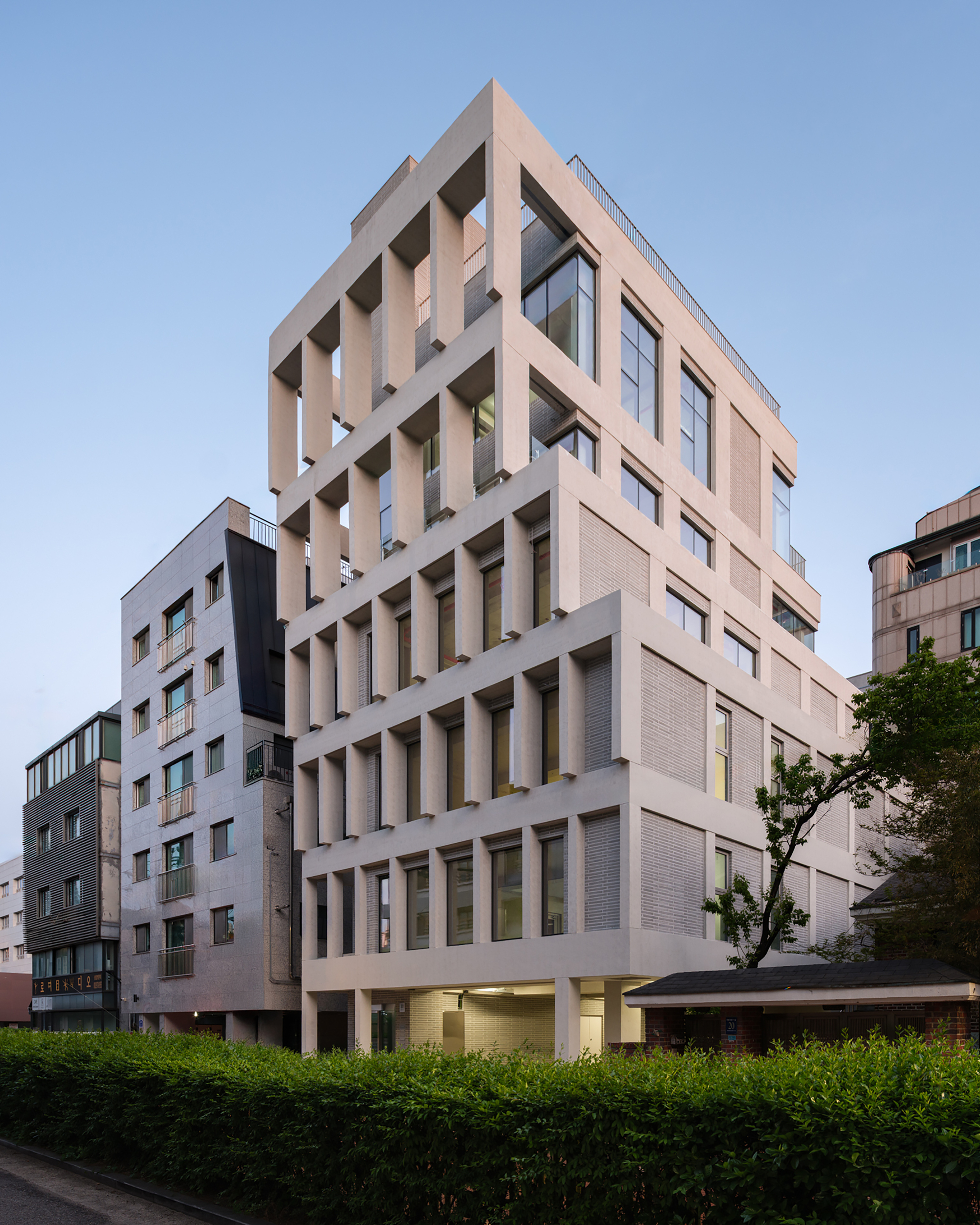 Corner view of the stepped concrete frame facade rising above a clipped hedge at dusk, with a leafy tree at right