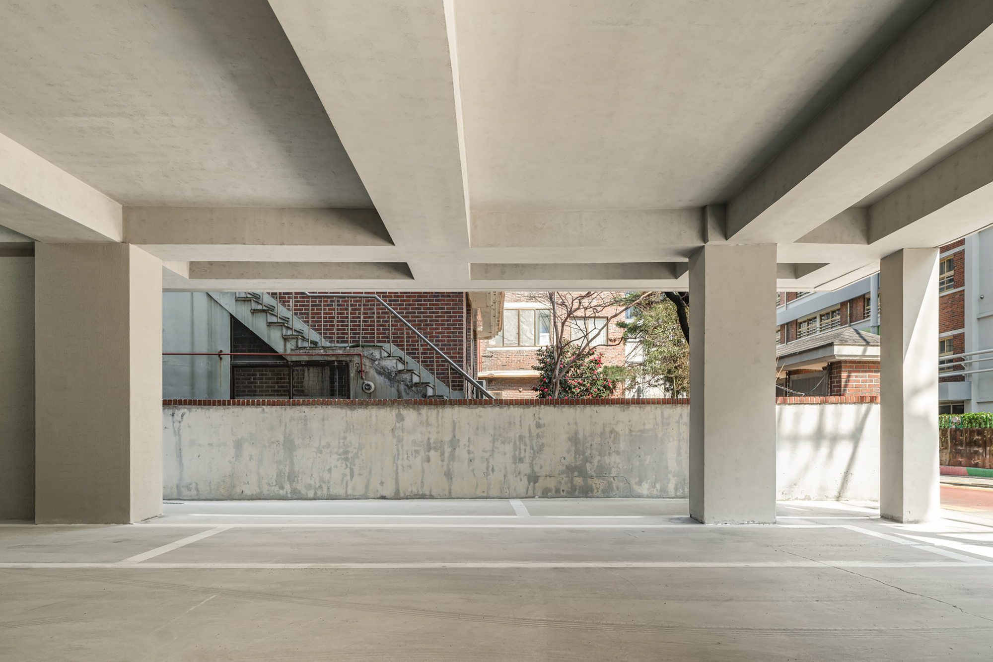 Open ground-level parking beneath concrete beams and square columns, looking out to a brick stair and neighboring houses in daylight
