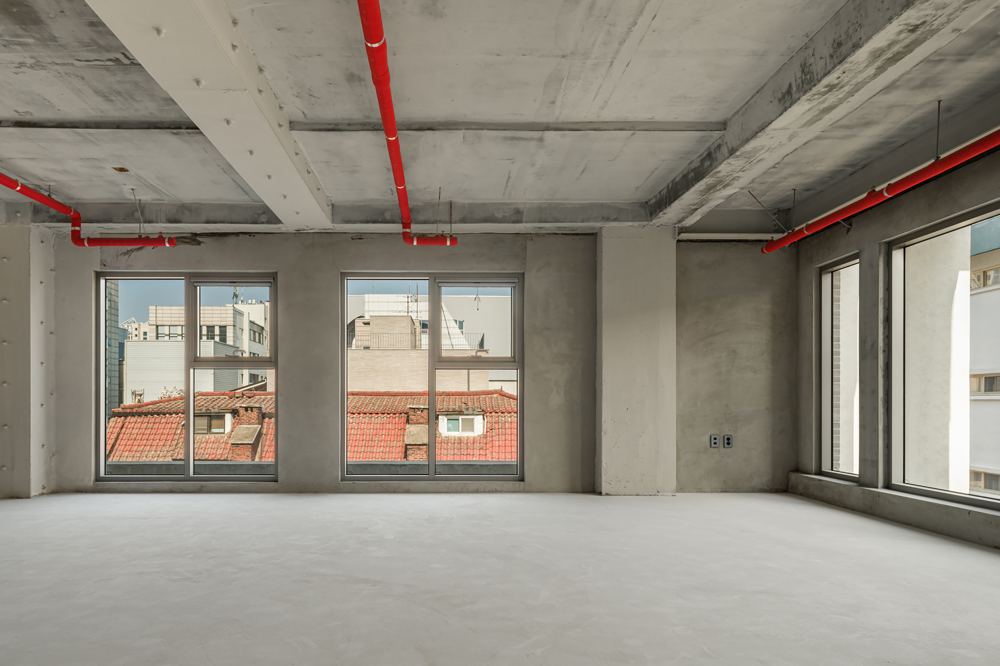Unfinished concrete room with red sprinkler pipes and tall windows facing red-tiled rooftops under a hazy sky