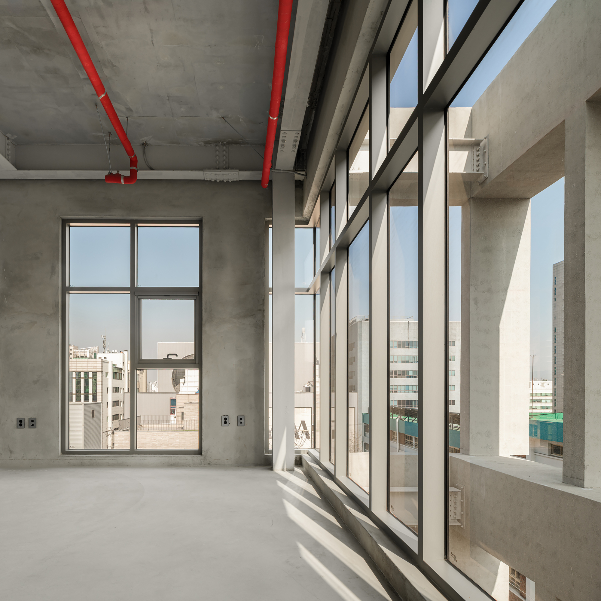 Corner of a raw concrete floor with a full-height glazed wall casting diagonal sunlight across the slab