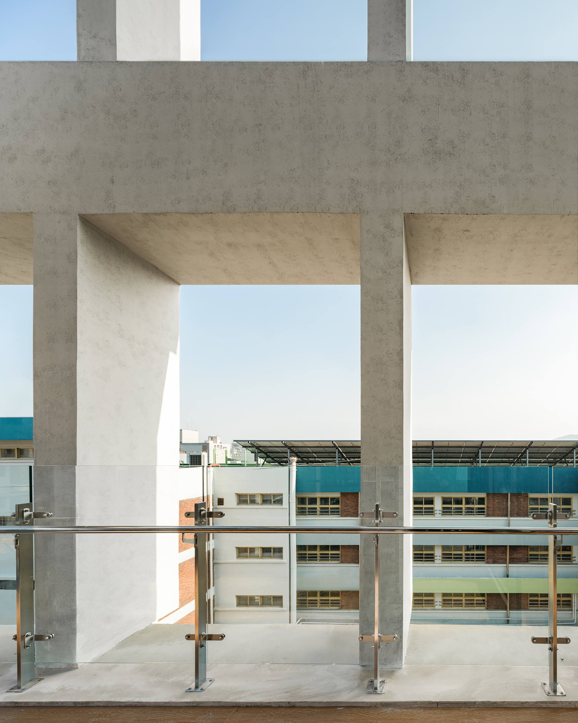 Concrete portal frame at a terrace with a steel and glass railing, looking toward a neighboring school block in clear daylight