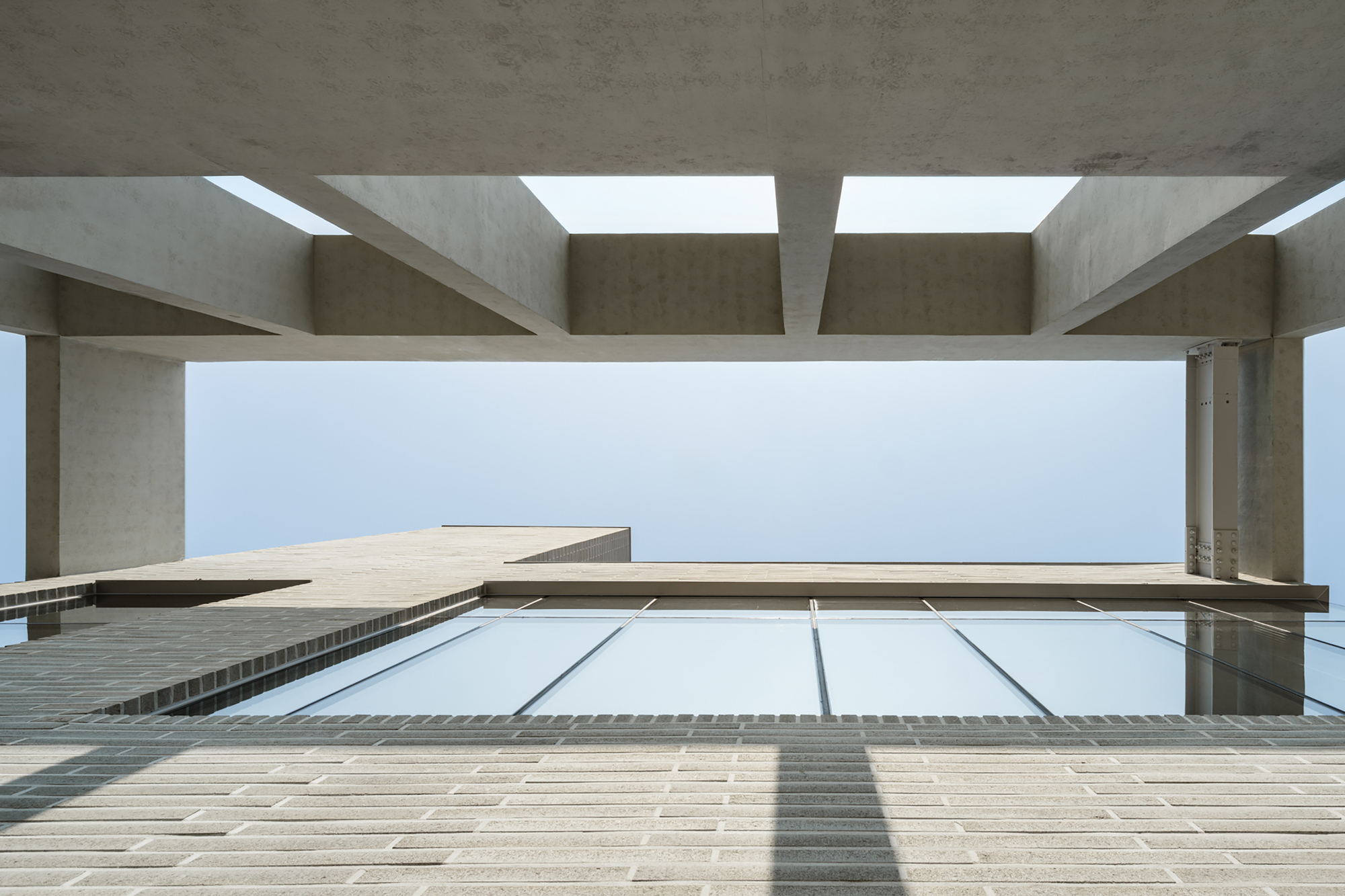 Upward view of the concrete beam grid above a brick paved terrace and sloped glass skylight against a pale sky