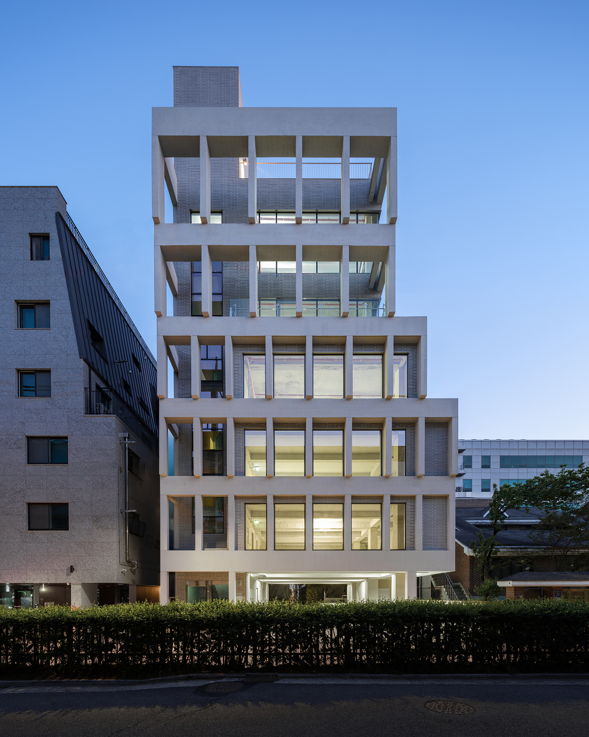 Street facade of the stepped concrete frame tower with lit floors at dusk, a clipped hedge in the foreground
