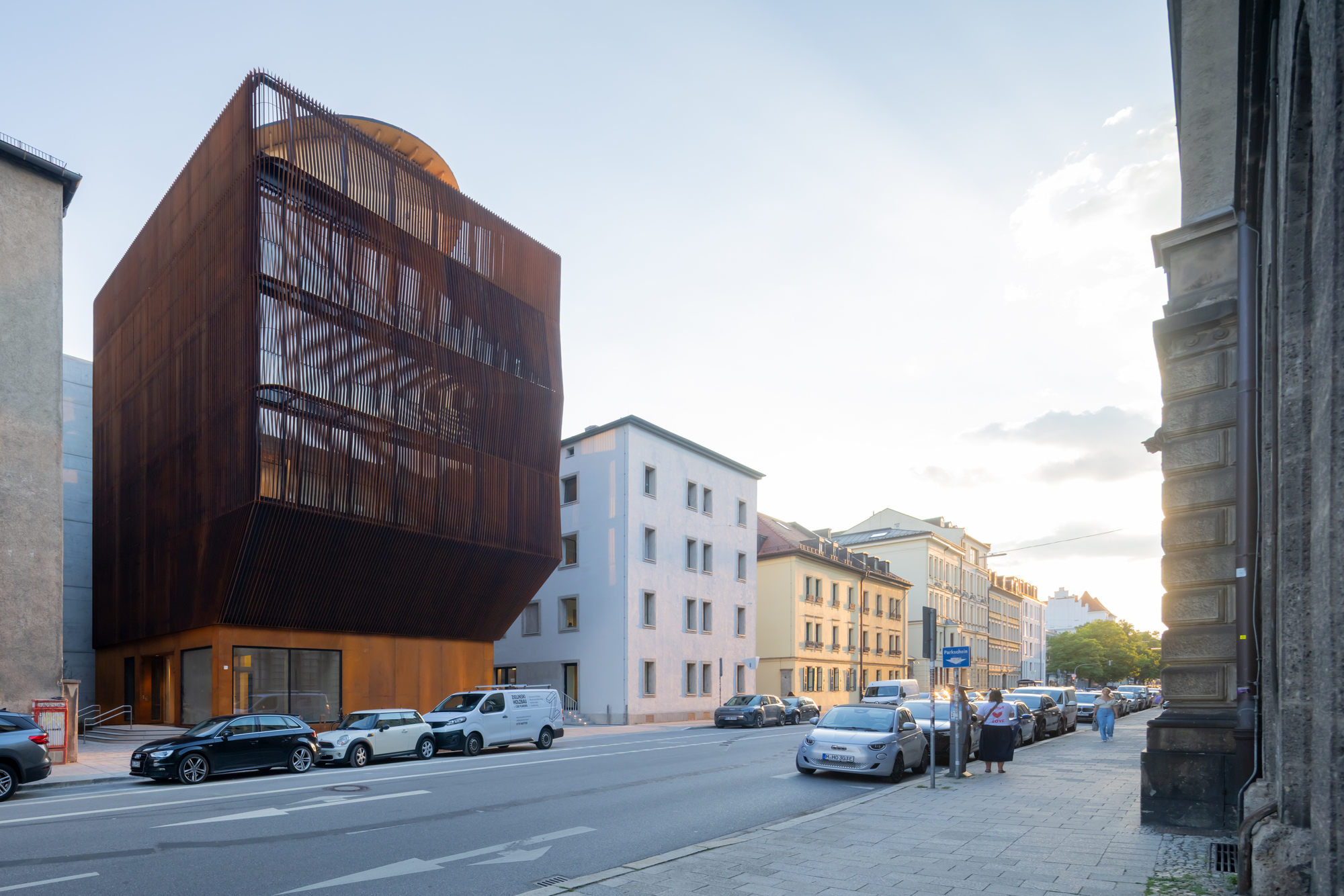 Corner view of the weathering steel slatted facade above a glazed base, with parked cars and pedestrians in warm evening light