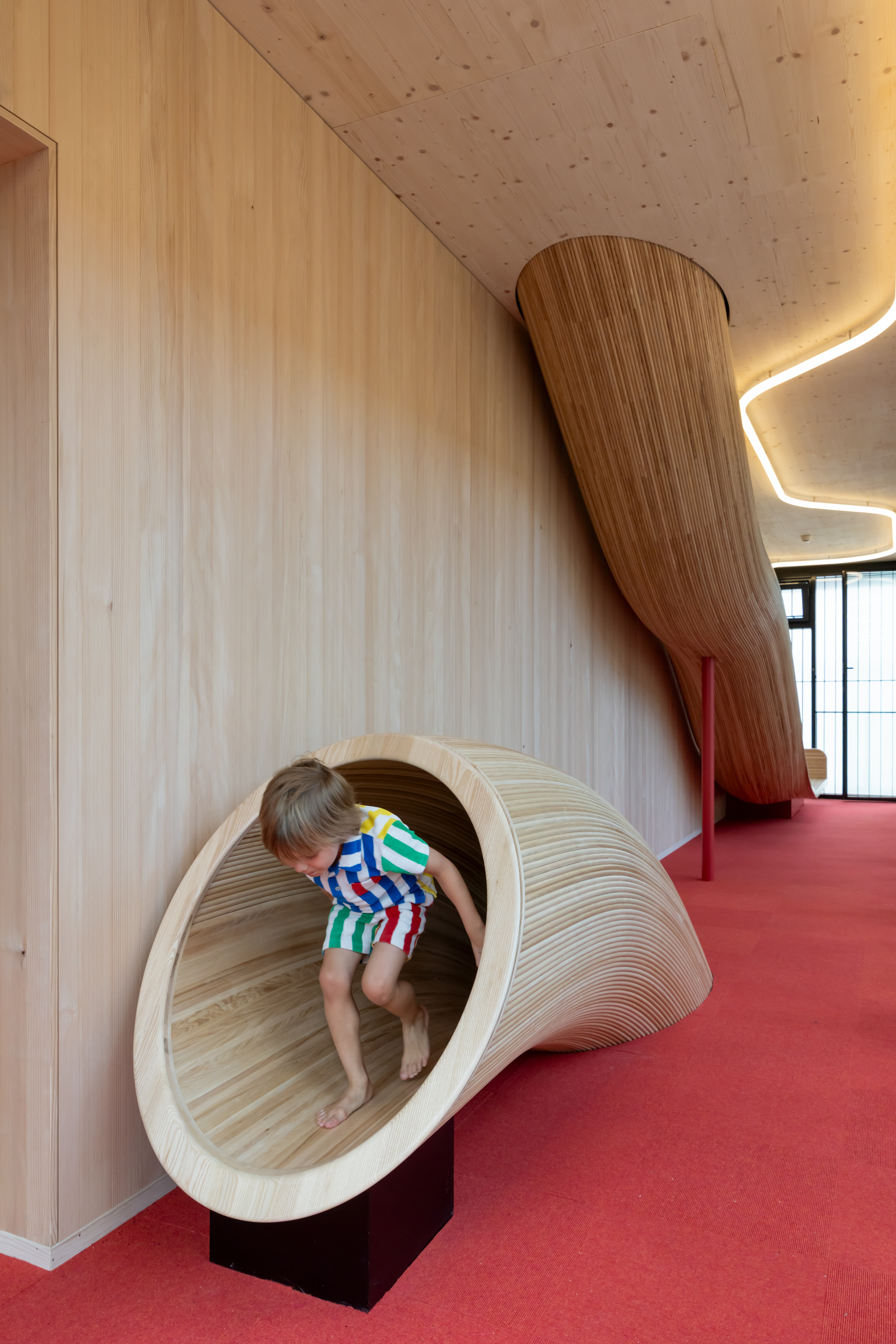 Child climbing out of a curved timber tube set against a wood-panelled wall beside a red carpeted corridor