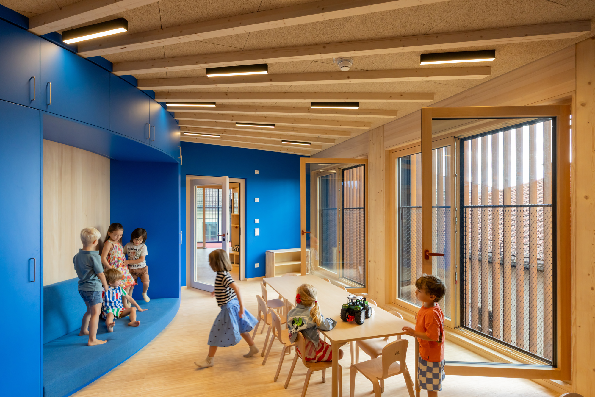 Classroom with a curved blue built-in bench, timber beams and tall wood-framed windows, with children playing at a table