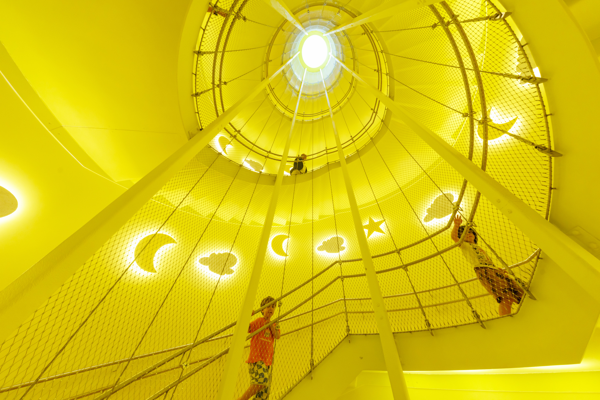 Upward view of a yellow stairwell wrapped in rope netting with glowing moon and star cutouts and children climbing