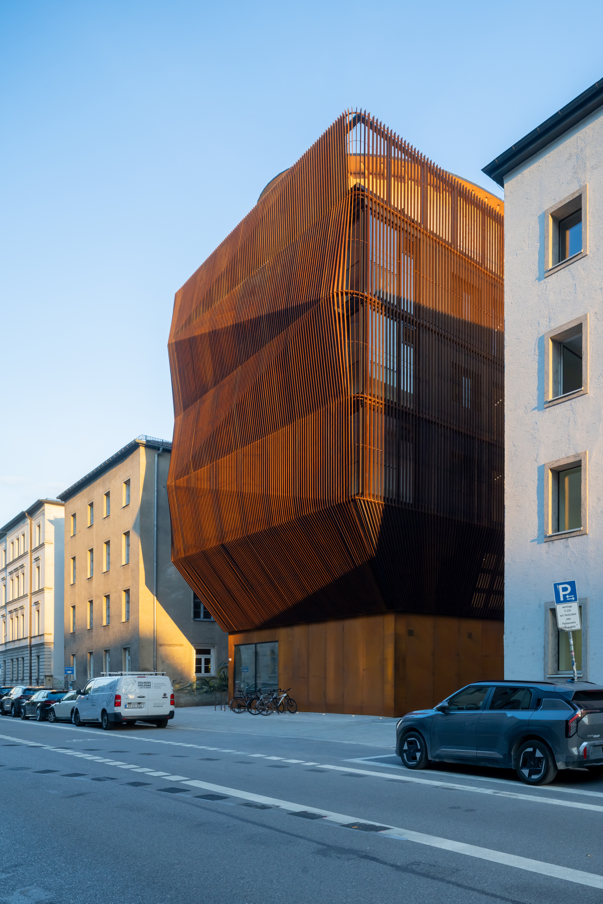 Faceted rust-coloured slatted volume rising over a copper-toned base, with a parked car and bicycles in low sun
