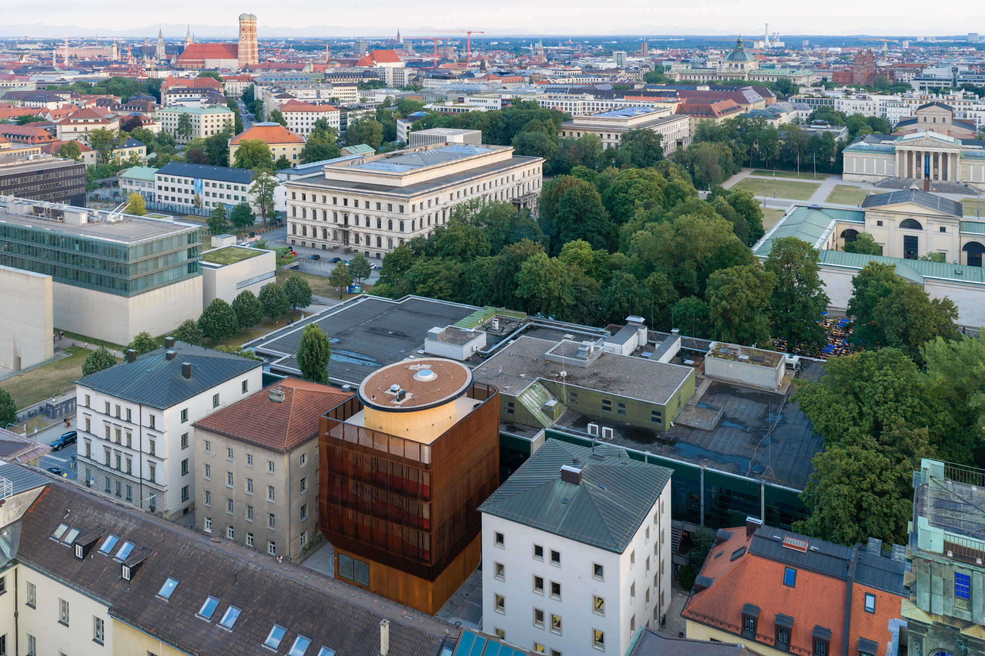 Aerial view of the rust-clad building with its round rooftop among older rooftops, green trees and distant towers under hazy sky