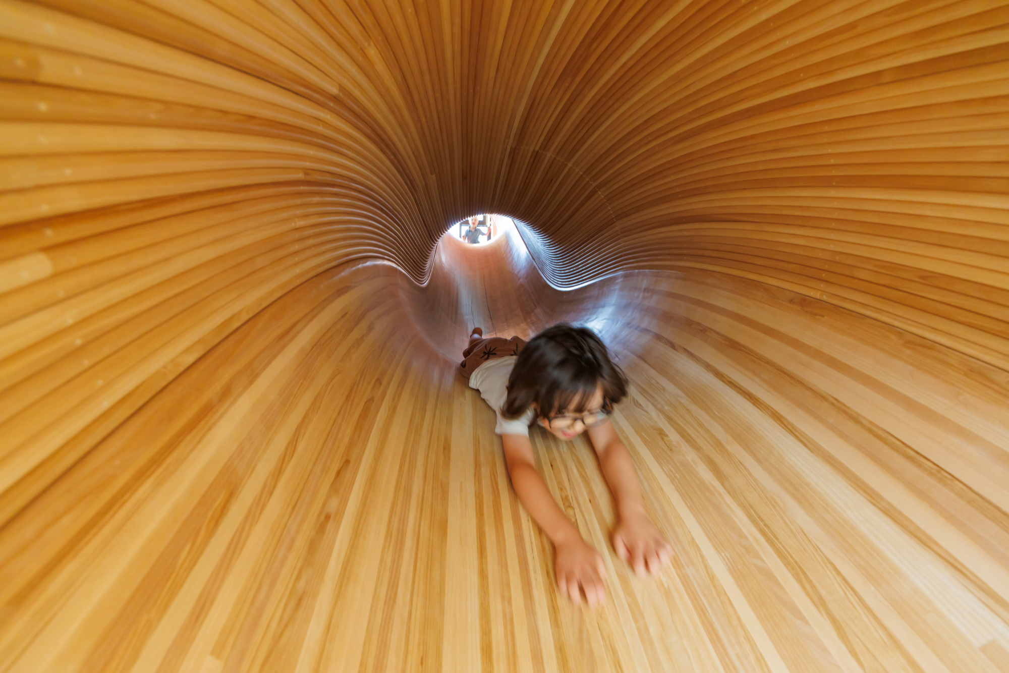 Child lying on the floor of a curved timber slide tunnel lined with thin wooden slats, another figure visible at the far opening