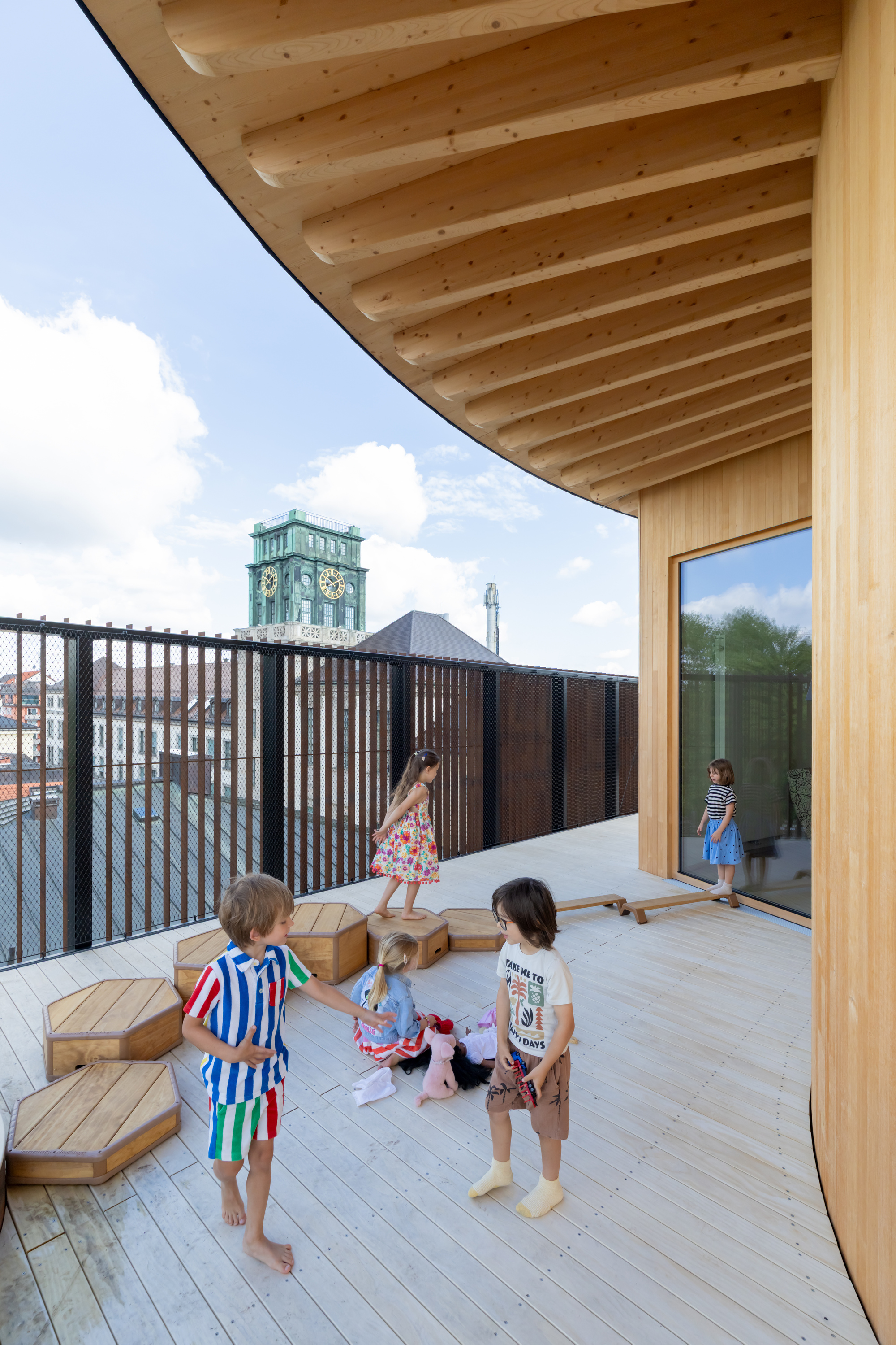 Children playing on a timber roof terrace with hexagonal stools, curved wall, exposed beams and a green clock tower under cloudy sky