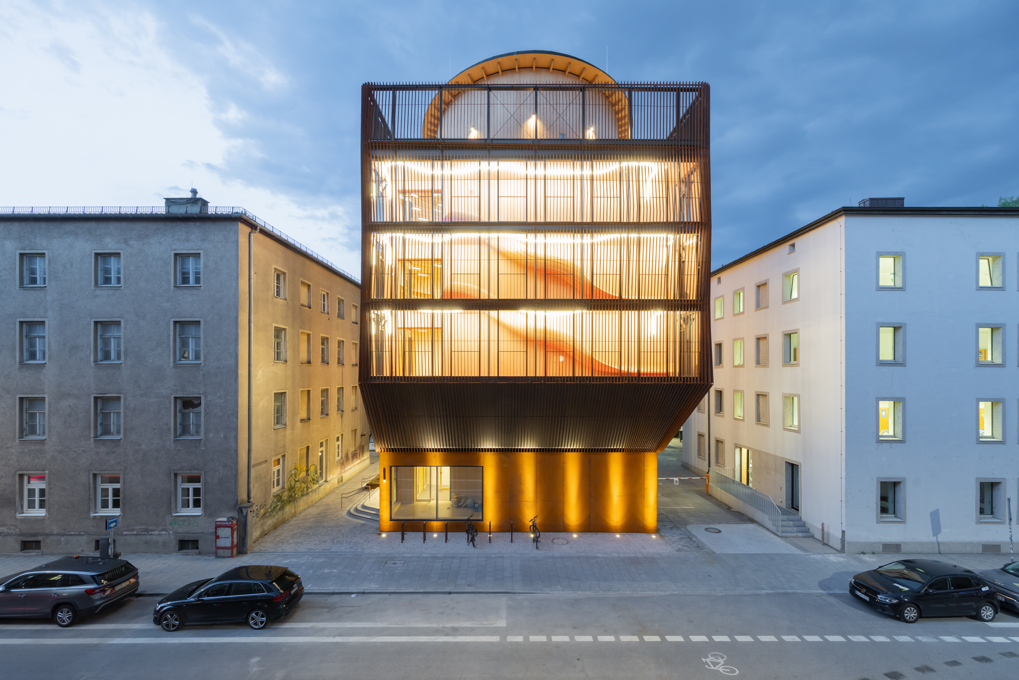 Street elevation at dusk of a boxy slatted tower with glowing interior and a domed timber roof between two older buildings