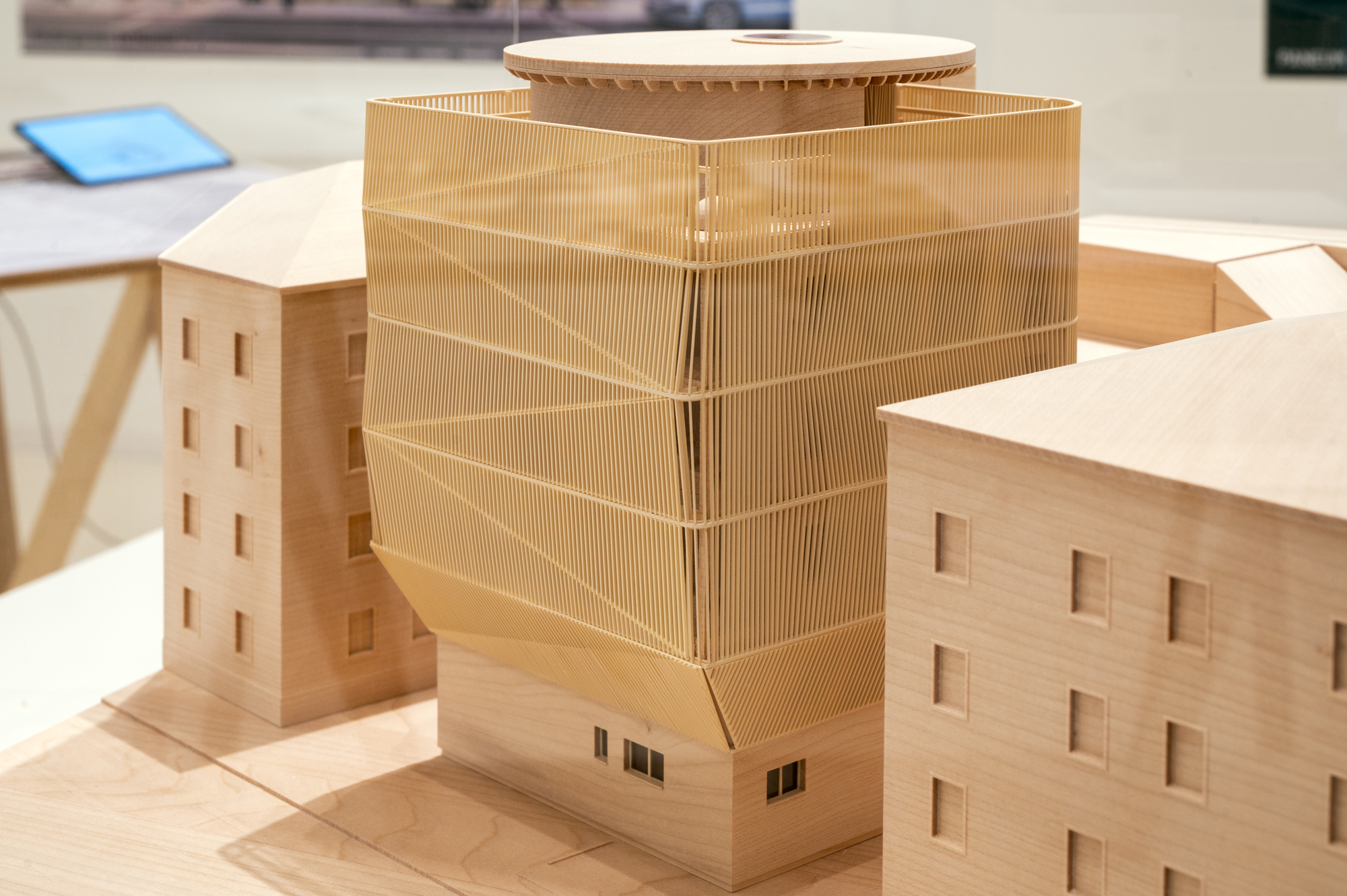 Wooden site model showing a faceted slatted tower with a round drum on top, set between two plain neighbouring blocks