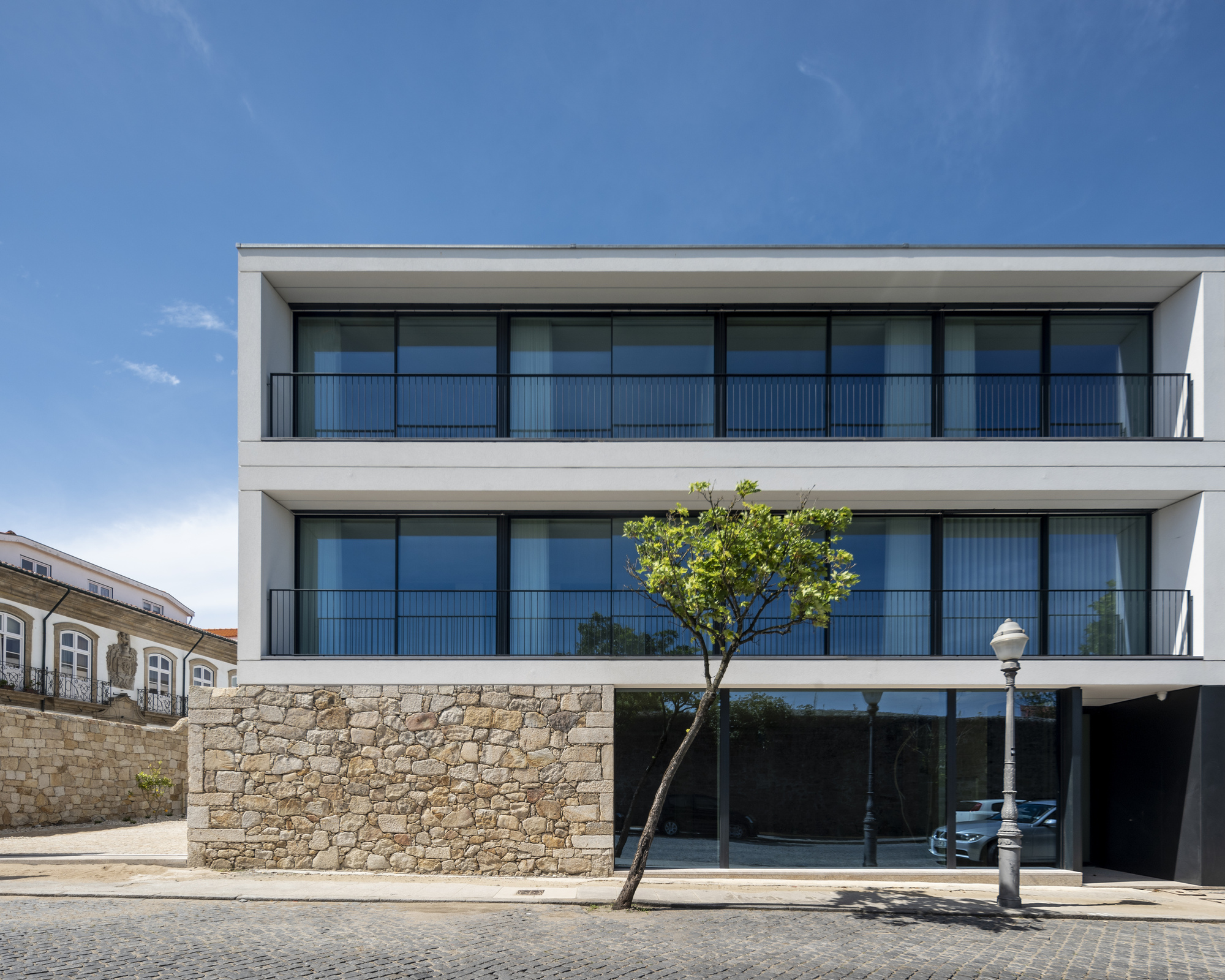 Three-storey white facade with black-framed glazing and railings above a granite rubble base, a young tree and lamp post in front under clear blue sky