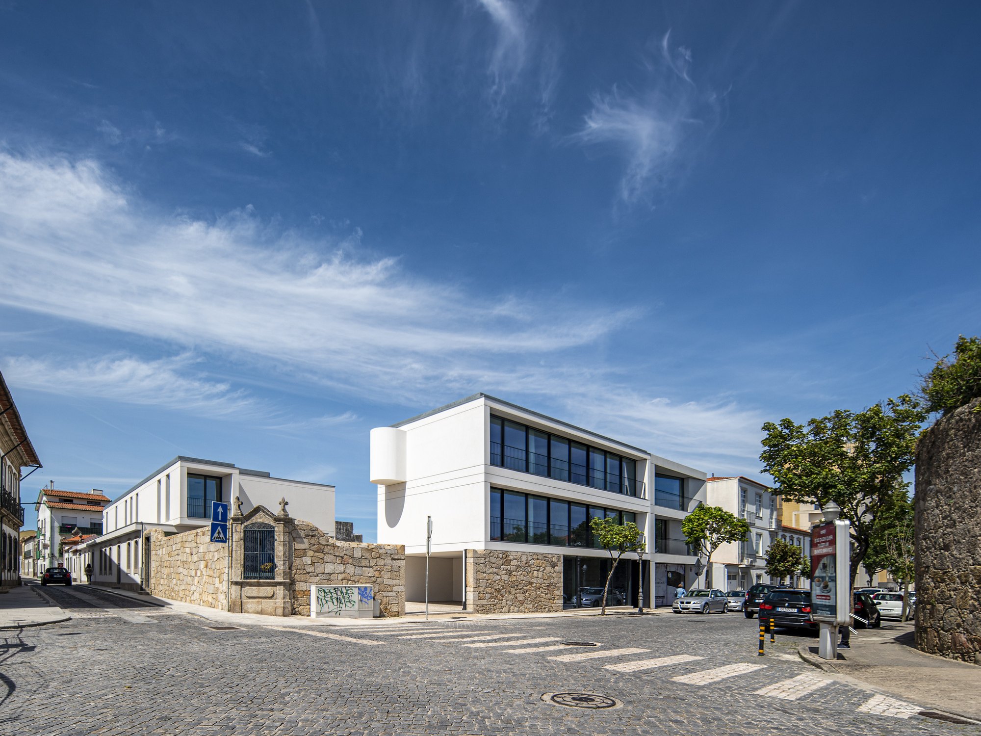 Street corner view of the white block with a rounded corner bay beside a granite wall, under wispy clouds