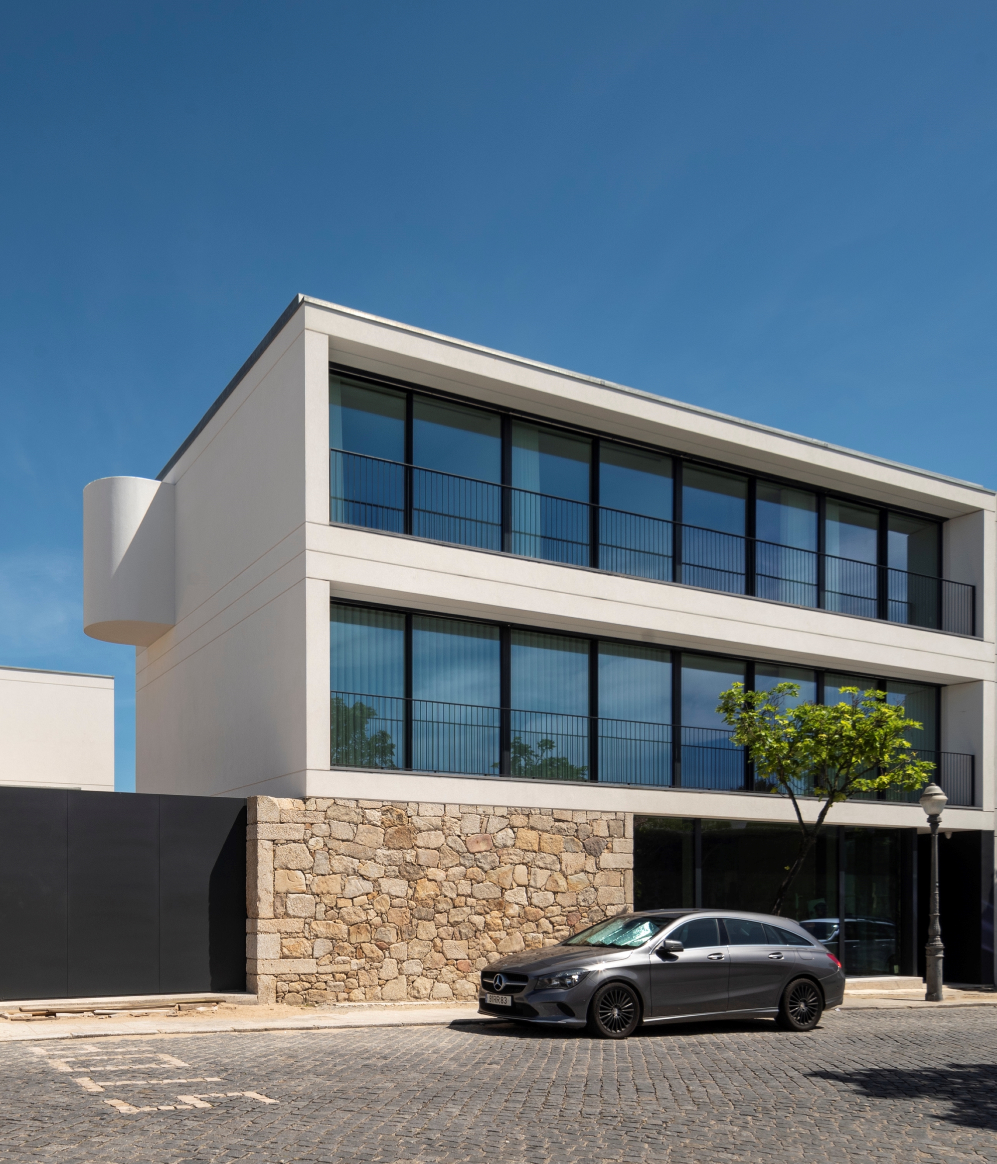Side view of the white upper floors and cylindrical bay over a granite base, with a grey car parked on cobbles