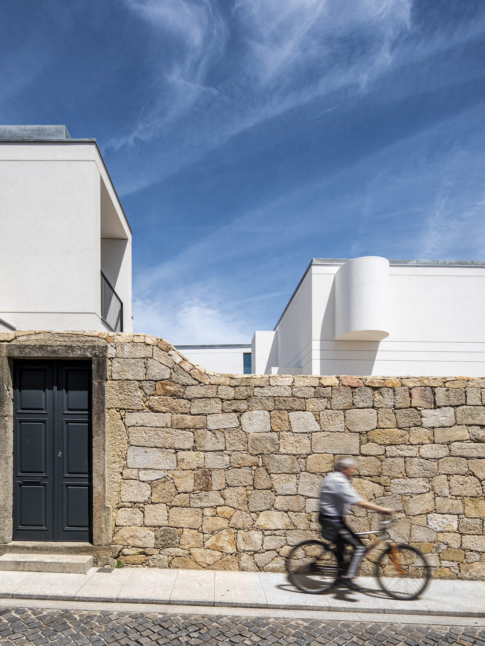 Granite rubble wall with a dark paneled door, white volumes rising behind and a cyclist blurred in motion on the pavement