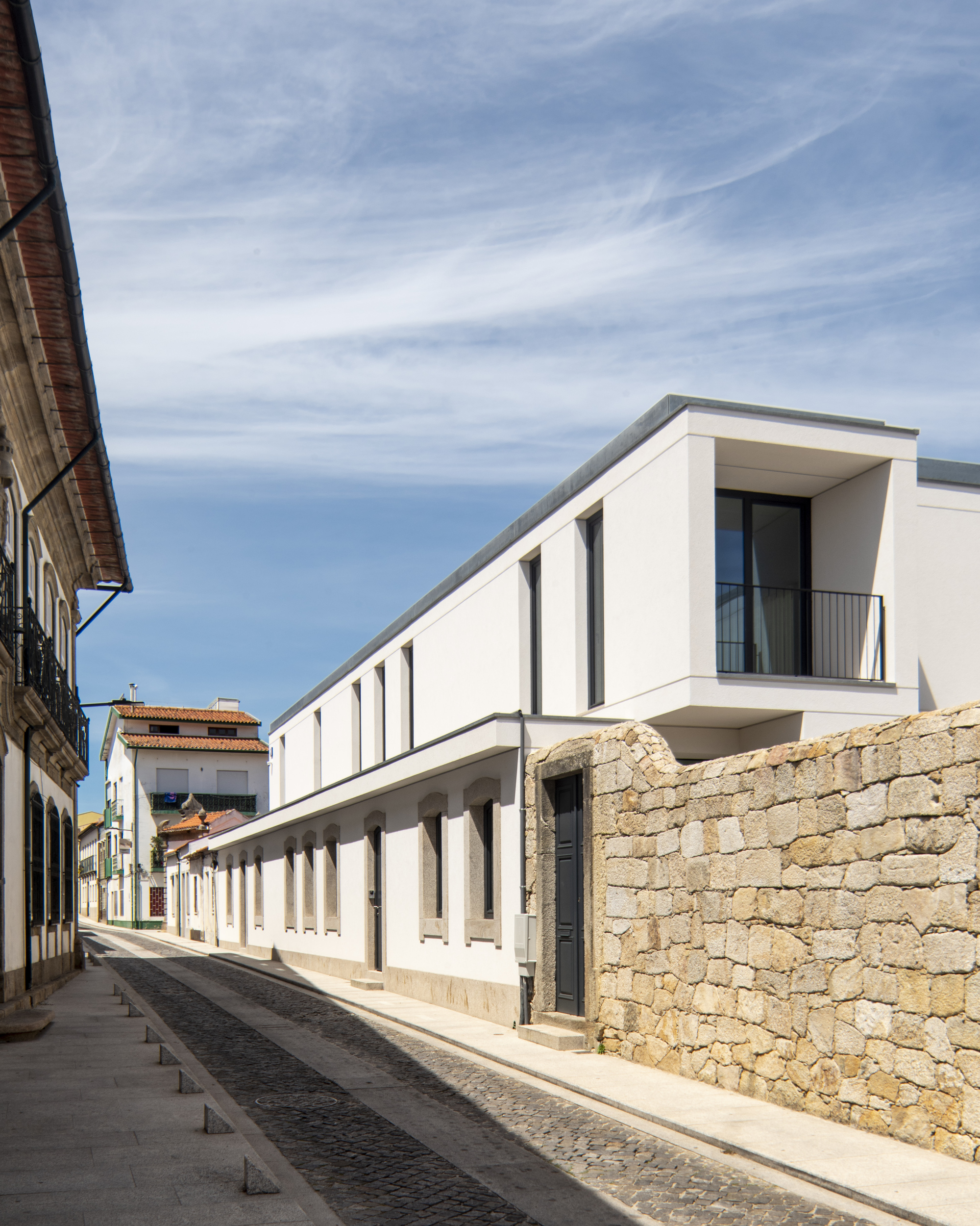 Narrow cobbled street running along a white elevation with a recessed balcony and a granite wall, beneath streaky cirrus clouds