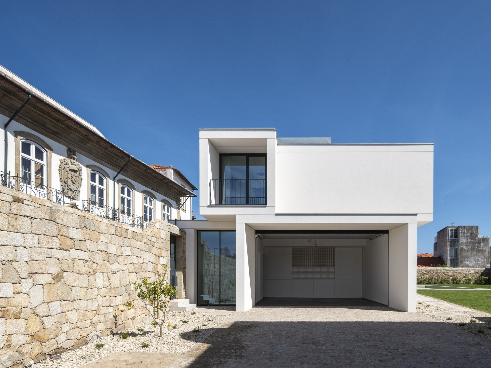 Rear courtyard view of the white cantilevered volume over an open carport, with an old granite wall and lemon tree in sunlight