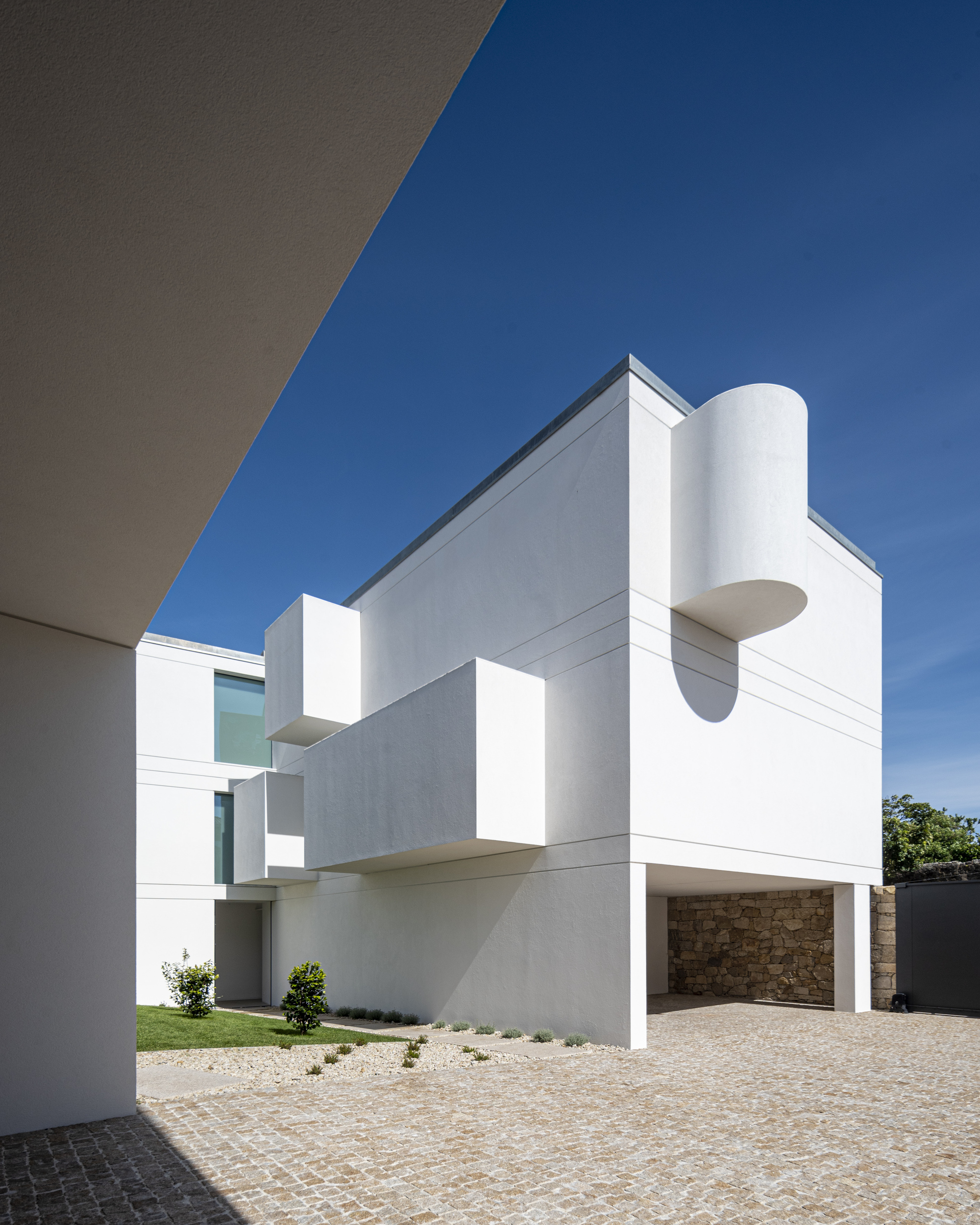 White stucco volumes with projecting boxes and a rounded drum above a granite-clad carport, seen across cobbled paving under deep blue sky