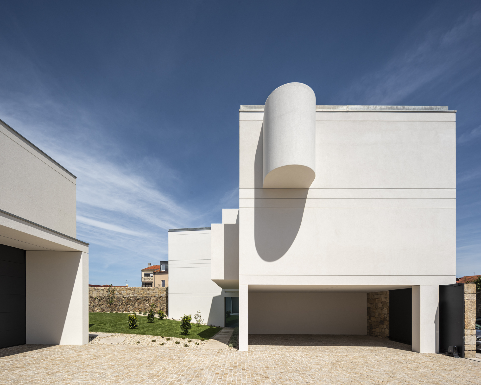 Front elevation of the white block with a cylindrical bay casting a long shadow over an open carport and sunlit cobbles