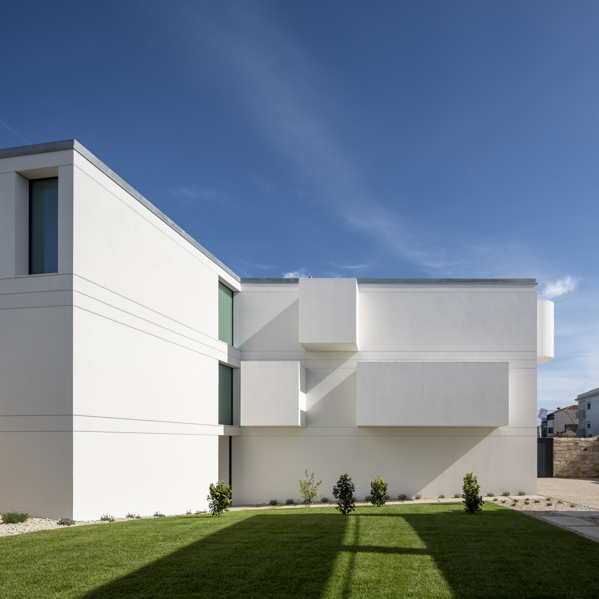 Side facade with staggered box projections above a lawn, trimmed shrubs and a long tree shadow in afternoon sun