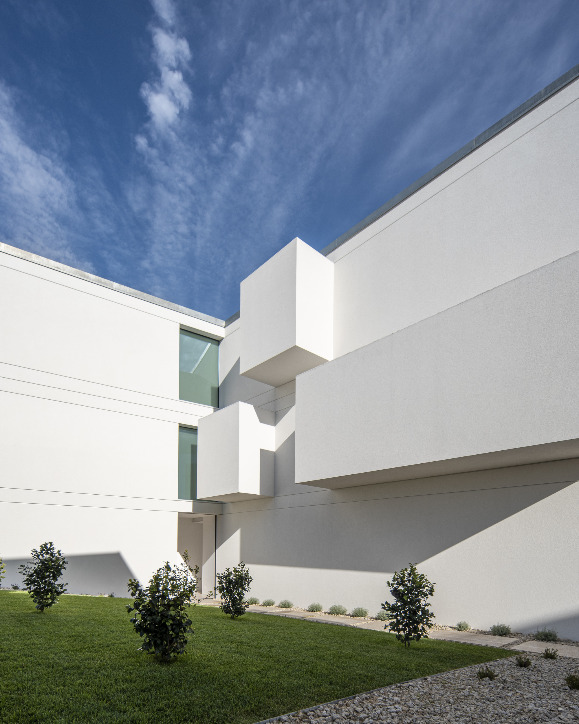 Low angle view of cantilevered white boxes and green-tinted windows above young trees and gravel, with wispy clouds overhead