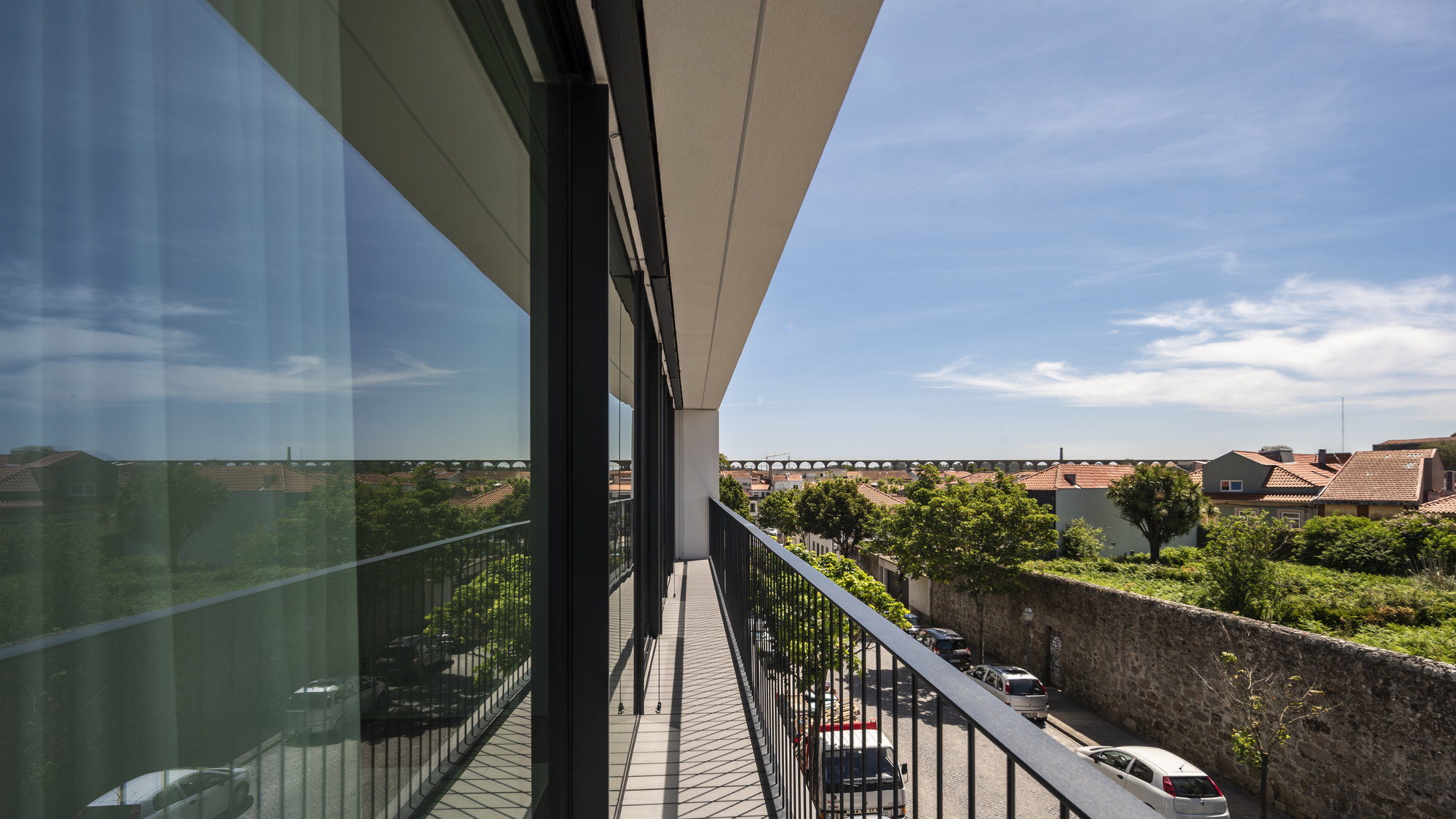Balcony view along a glazed wall with black railing, looking over a stone wall, parked cars and a distant aqueduct