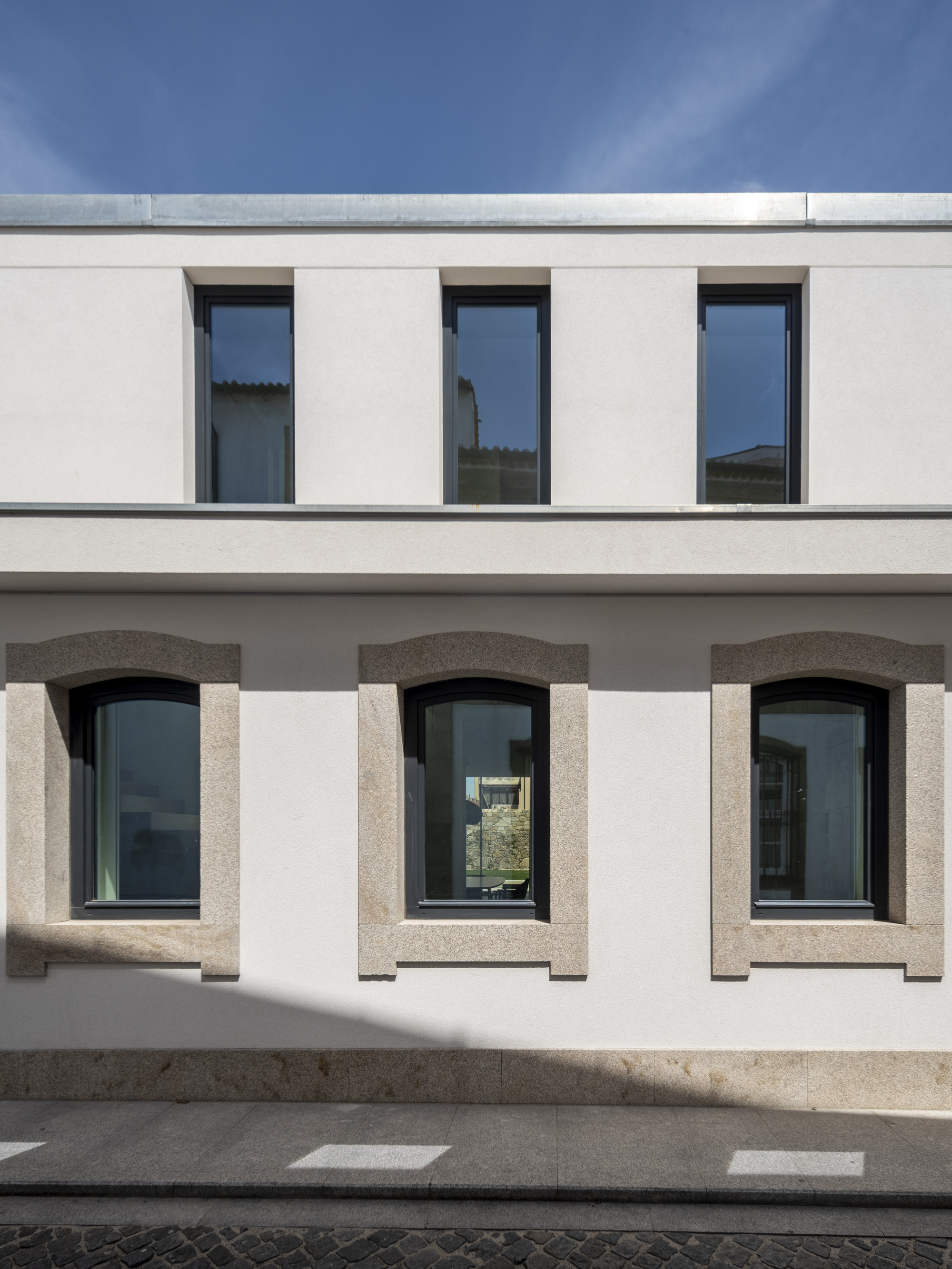 Rendered white facade with granite-framed arched windows below tall rectangular openings, with sharp shadow falling across the paved footpath