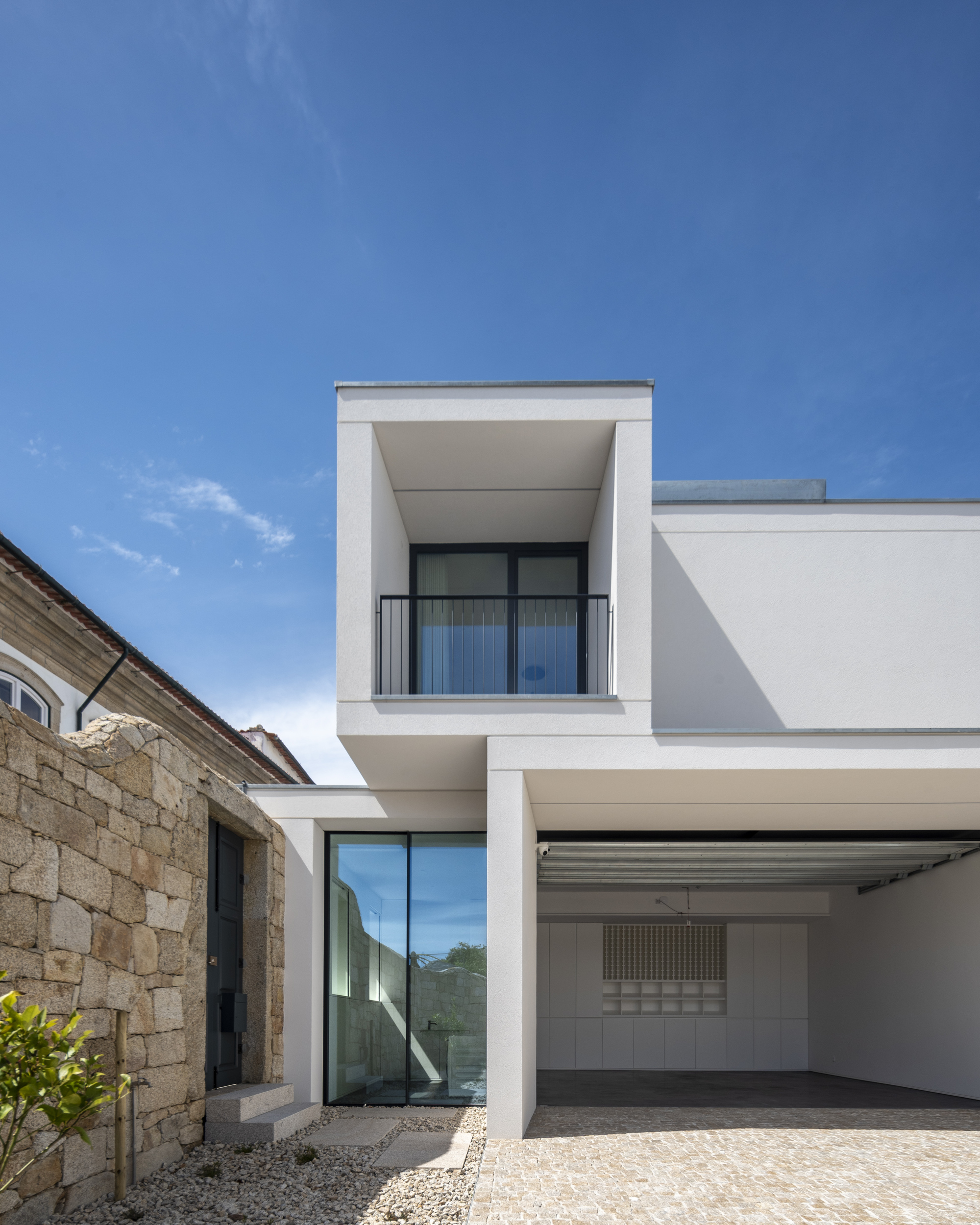 Two-storey white volume with a recessed balcony and open carport beside a rough granite wall under a clear blue sky