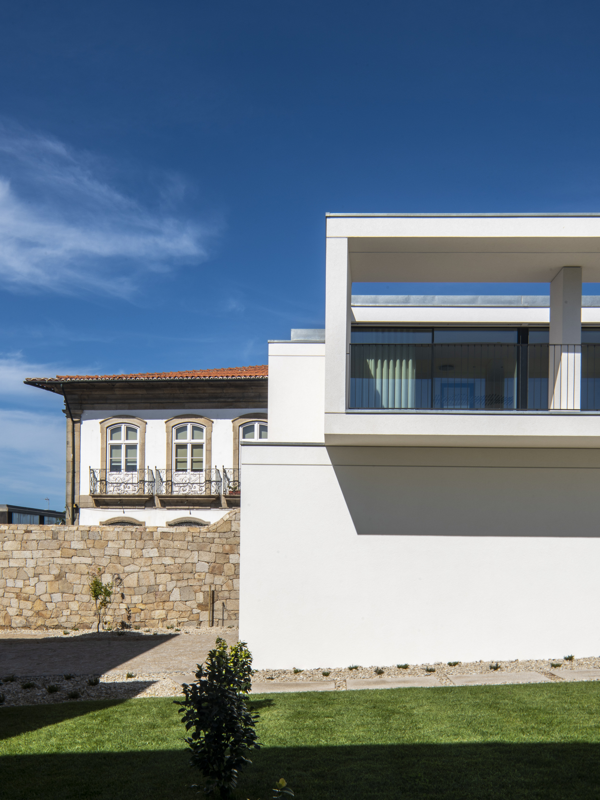 White upper terrace with slim columns and glass guardrail beside an old stone wall and tiled-roof house, seen over a lawn