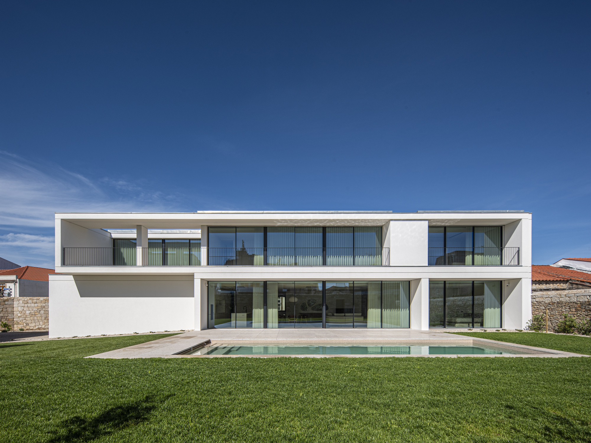 Rear elevation of the long white house with glazed ground floor and a narrow pool across a mown lawn in bright sun