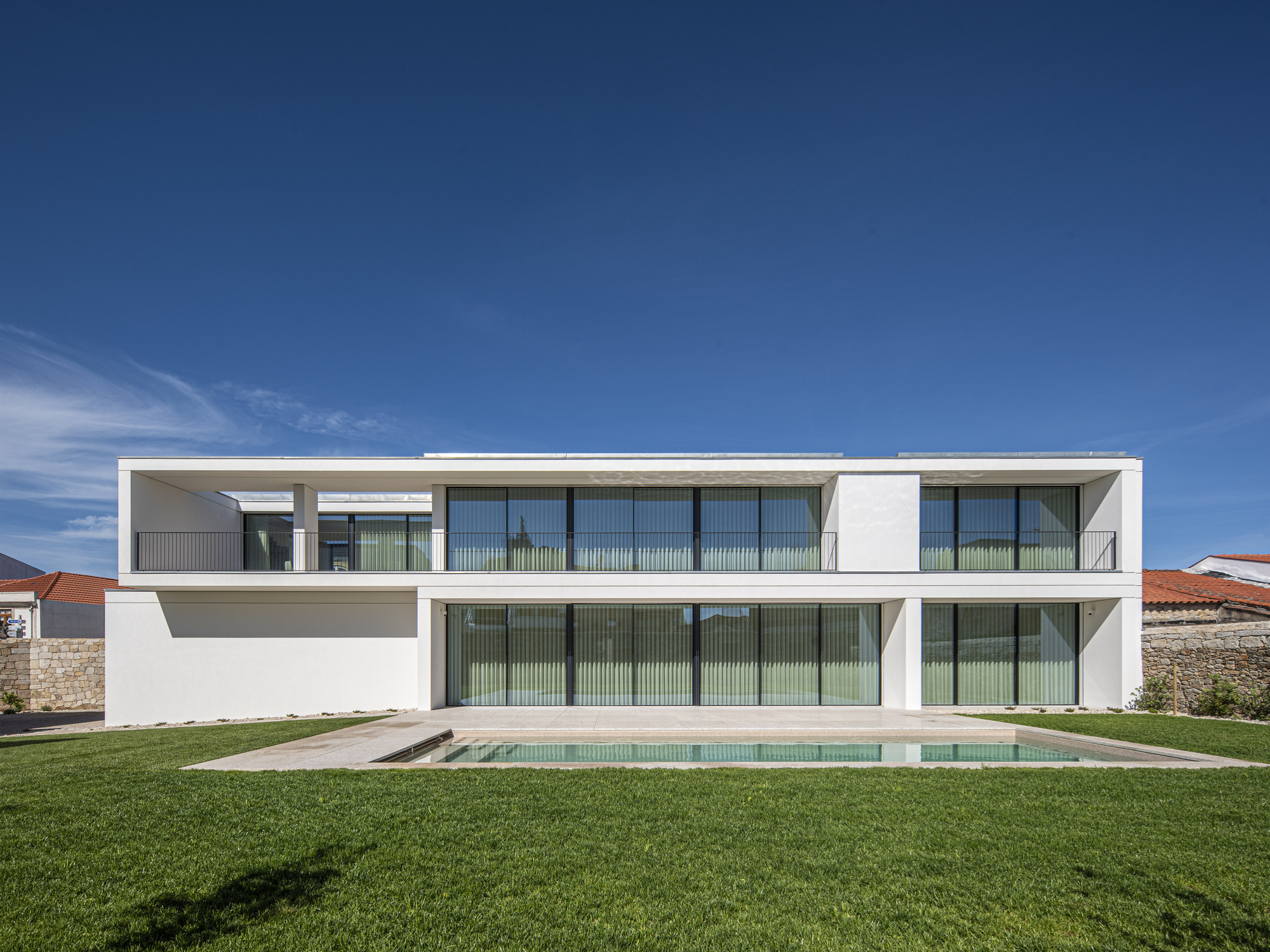 Rear elevation with curtains drawn behind the full-height glazing, the pool and lawn in the foreground beneath a deep blue sky