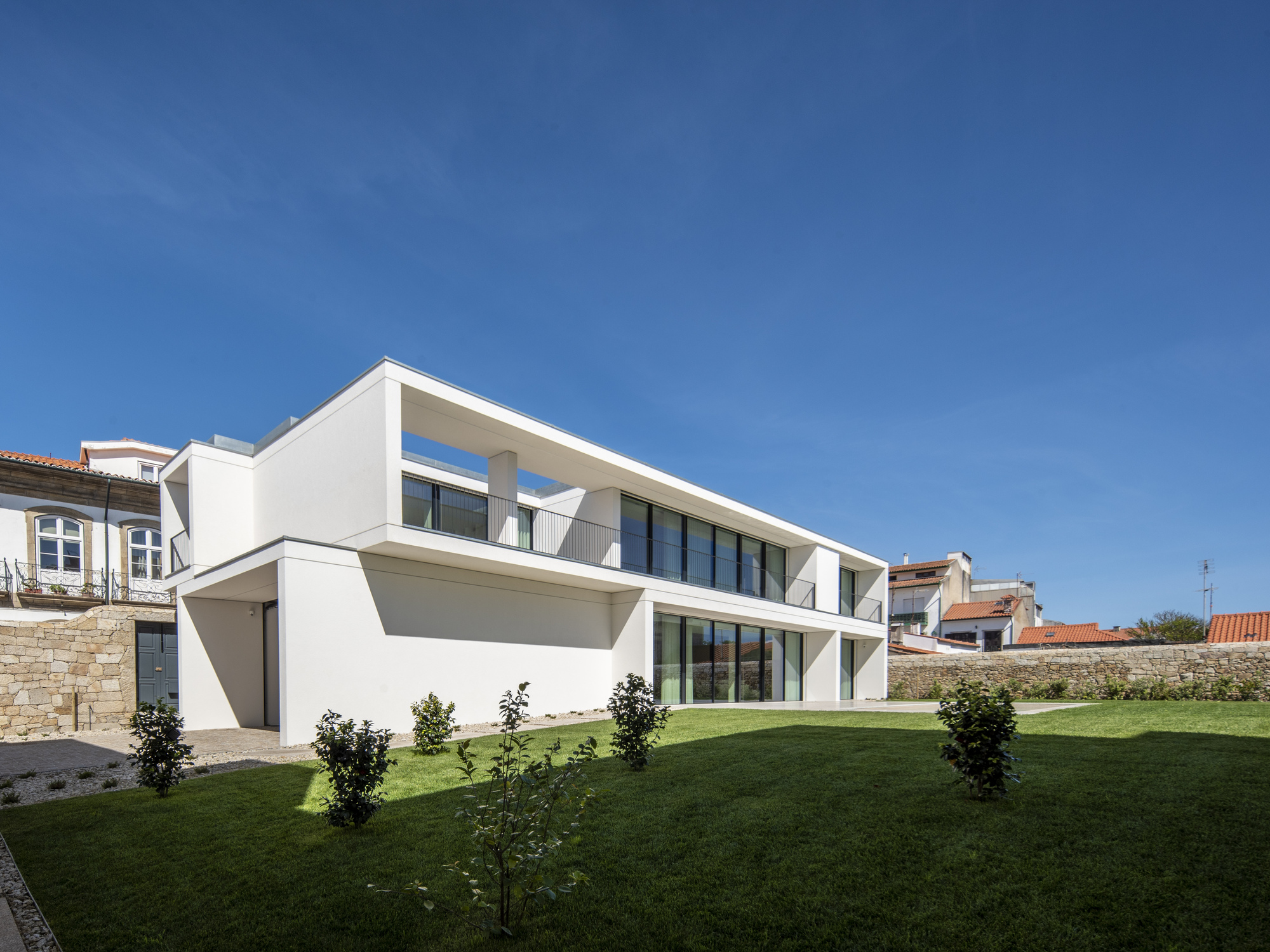 White two-storey volume with a cantilevered upper floor and glazed ground level, seen across a lawn with young shrubs under a clear blue sky
