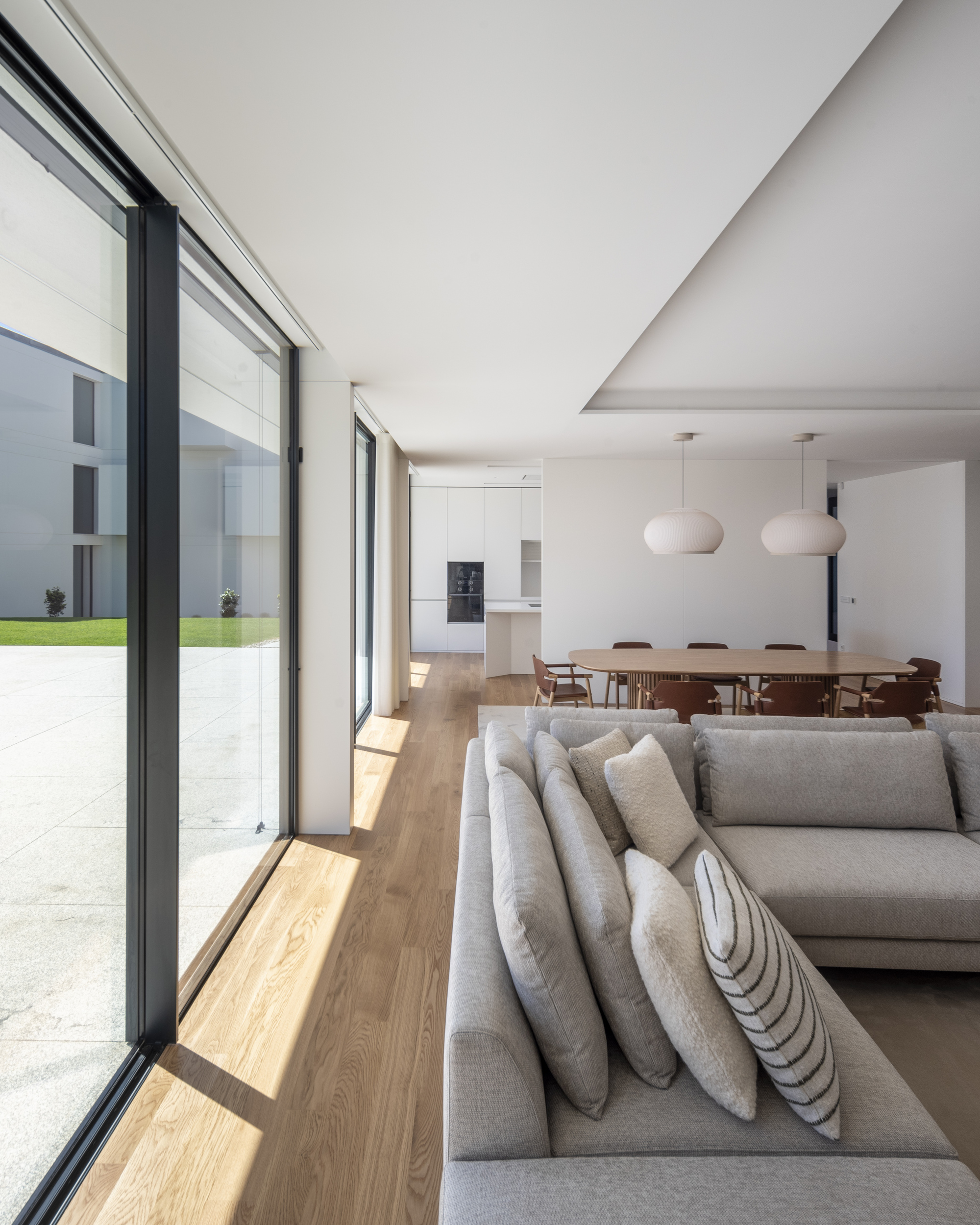 Living room seen past a grey corner sofa toward the dining table, with sunlight falling across the oak floor from a sliding glass wall