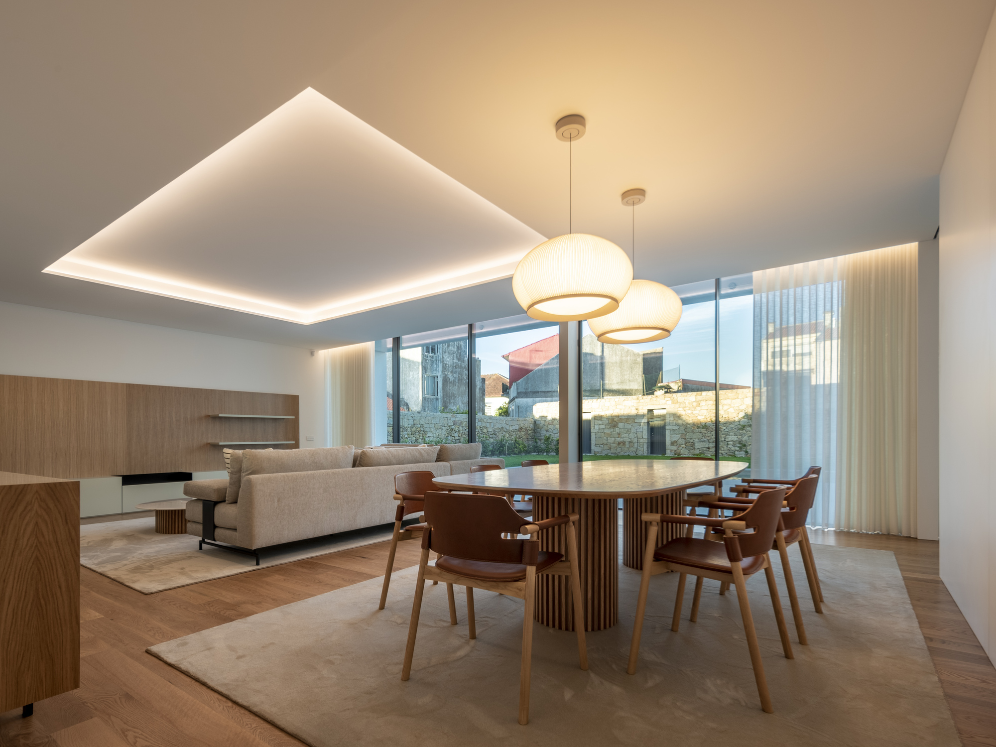 Dining table with leather chairs and two pendant lamps beneath a lit recessed ceiling panel, with a stone garden wall visible through the glass