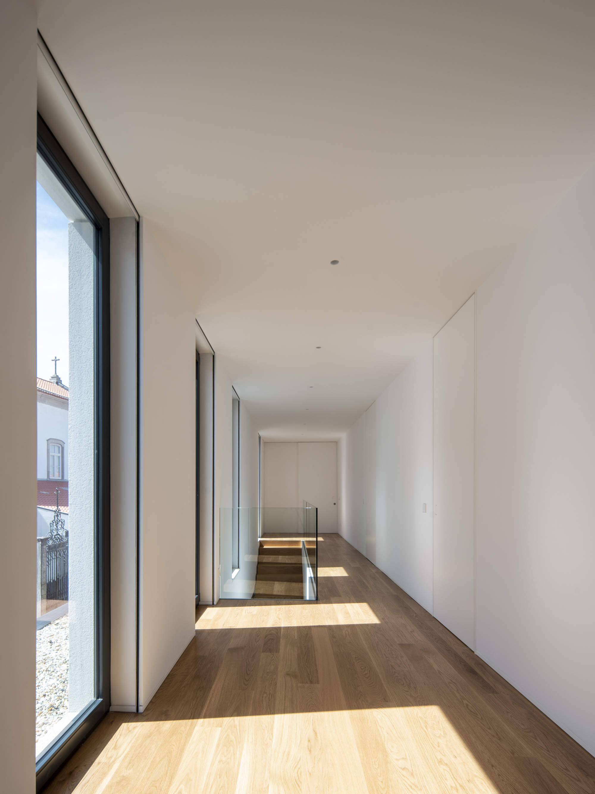 Upstairs corridor with oak flooring, a tall window on one side and a glass stair balustrade at the far end, lit by afternoon sun